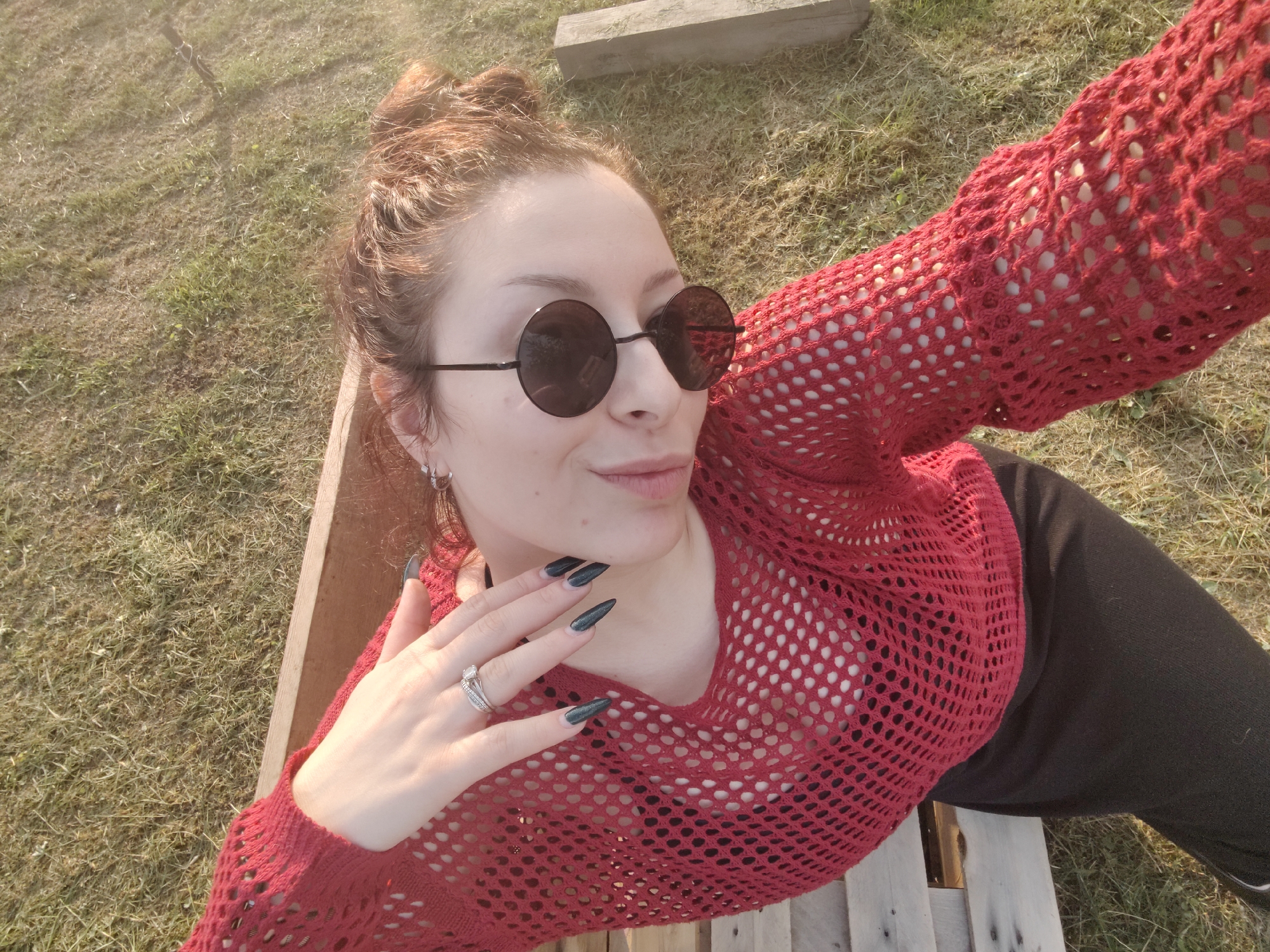 In this outdoor, selfie-style photograph, a young woman with auburn hair styled in two buns is seen lying down, possibly on a wooden pallet or bench. She is wearing round, dark sunglasses and a red, mesh, long-sleeved top over a dark undergarment. Her left hand, adorned with a silver ring and long, pointed, dark green nails, is raised to her cheek.
The scene is set on a grassy area with dry patches, suggesting a sunny day. A wooden plank is visible in the background, and another wooden structure, possibly a pallet, is beneath the woman. The lighting indicates it is late afternoon or early evening, with warm, golden sunlight casting shadows. The woman's expression is relaxed and perhaps playful, with her lips slightly parted and her gaze directed towards the camera.
The image captures a casual, leisurely moment outdoors, likely during a warm season. No visible text is present in the photograph. The location context provided is Ермолаево, Russia.