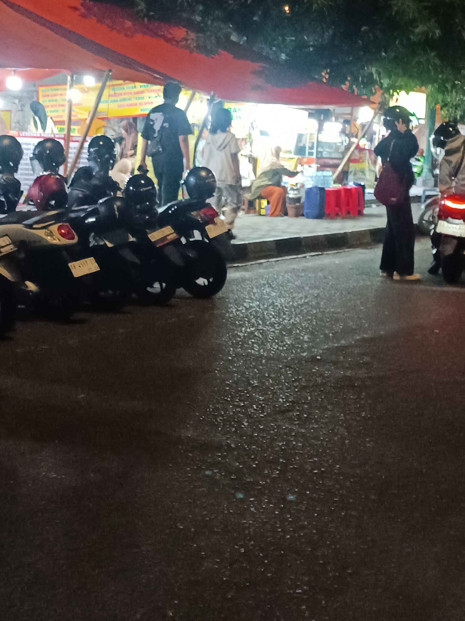 The image depicts a nighttime scene at an outdoor food stall area in Yogyakarta, Indonesia, likely on a street. Several motorbikes are parked in the foreground. In the background, a food stall with an orange canopy is visible, with lights illuminating the area. People are seen walking and standing around the stalls. One person is walking away from the camera, wearing a black t-shirt with a white graphic. Another person, wearing dark clothing and carrying a maroon bag, stands facing the stall. There are also individuals seated and standing further within the stall area, obscured by the lighting. The ground appears wet, suggesting recent rain. The overall atmosphere is that of a lively, urban street food scene at night.