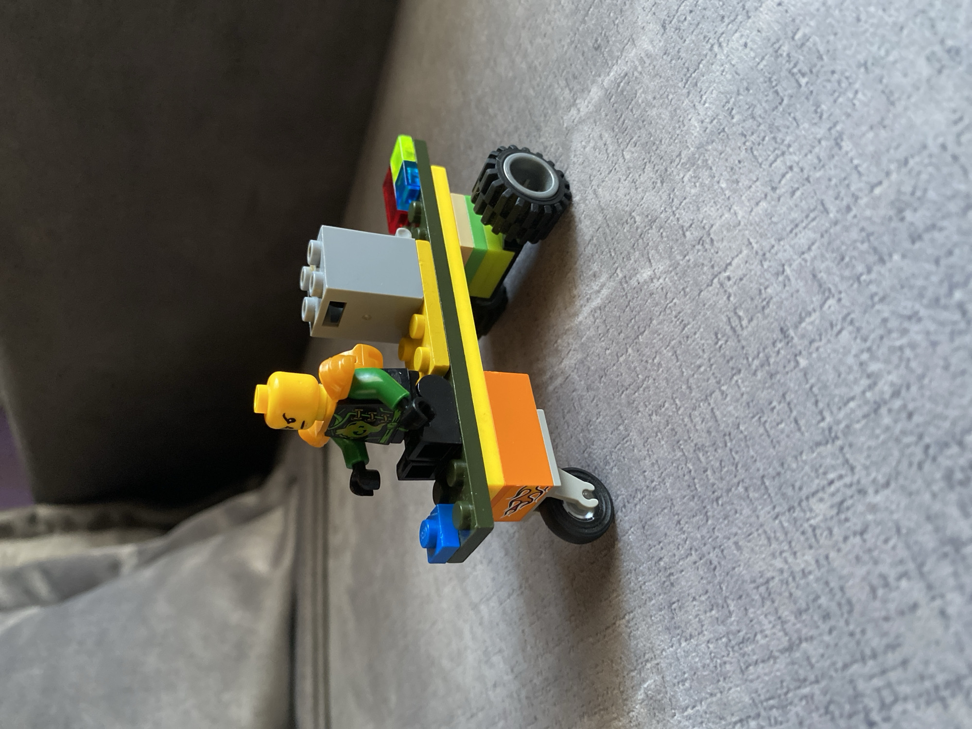 A Lego minifigure with a yellow head, green torso, and orange shoulder pads is seated on a Lego vehicle. The vehicle has a yellow minifigure torso-like piece as its front, with orange and yellow blocks forming its body. It has two wheels at the back and one smaller wheel at the front. The minifigure is holding onto the vehicle, and there is a gray Lego brick block with two visible studs on the back of the vehicle. The Lego creation is placed on a gray textured surface, possibly a sofa or carpet. The background shows a soft, out-of-focus gray object, likely a cushion or furniture. The overall scene is a close-up indoor shot.