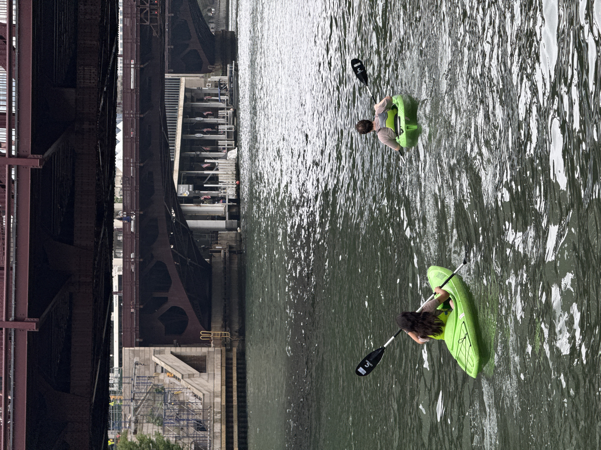 Two people are kayaking on the Chicago River, a waterway flowing through the city of Chicago, Illinois, United States of America. The kayaks are bright green. The person in the foreground, with long dark hair, is wearing a grey t-shirt and a yellow life vest. They are paddling a kayak with a black and white paddle. The second person, in the middle ground, is wearing a tan t-shirt and a grey and green life vest. They are also paddling a kayak with a black and white paddle. In the background, there are several bridges crossing the river. The closest bridge is a large, dark red metal structure. Below this bridge, there are buildings with flags hanging outside, and a boat docked near the riverbank. Further in the background, another, higher bridge is visible. The water of the river is a dark, murky green, with ripples and reflections from the overcast sky. The scene suggests a recreational activity taking place within the urban landscape of Chicago.