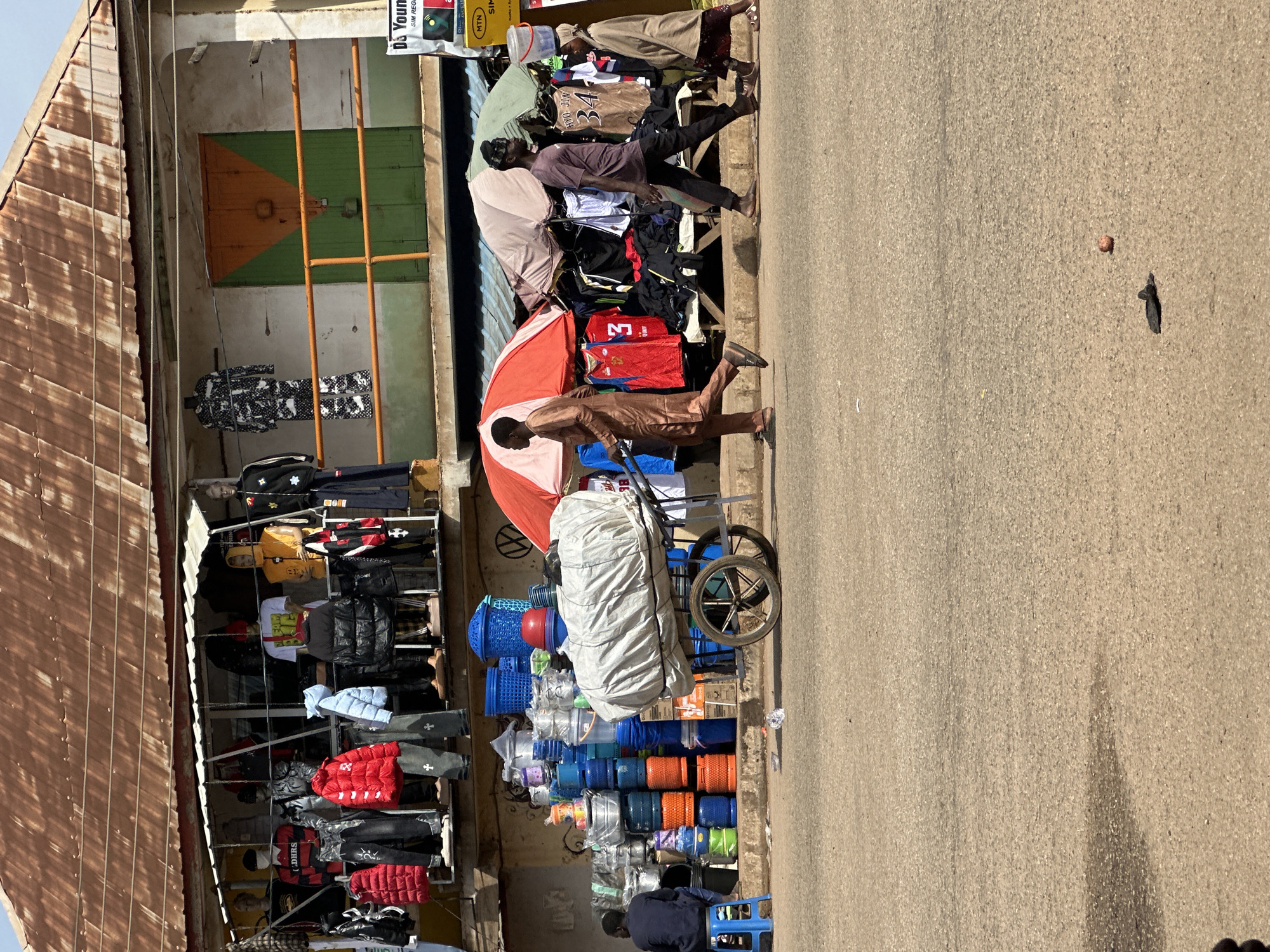 This is a street-level, medium shot photograph taken in Bukuru, Nigeria, depicting a bustling market scene under a corrugated iron roof. In the foreground, a paved road occupies the lower half of the image, with a shadow falling from the left. To the left of the road, a person in a dark blue shirt and pants is seated on a blue stool, facing away from the camera. Adjacent to them is a large stack of various plastic and metal containers, including buckets and pots, predominantly in shades of blue, orange, and silver. A two-wheeled cart with a large, light-colored bundle covered in fabric is positioned in the center, with one wheel prominently visible.
In the midground, several people are walking on the side of the road. On the left, a person in a light brown garment is walking away from the camera, carrying a long, thin object. In the center, under a red and white umbrella, two men are walking. One is wearing a dark purple shirt and dark pants, and the other is obscured. To the right of the umbrella, a woman in a light brown headscarf and patterned skirt is walking towards the right side of the frame. Behind the people and the cart, a market stall is visible, displaying an assortment of clothing on mannequins and hangers, including jackets, shirts, and pants. The stall has a metal railing in front of it and a green and orange door in the background. The building above the stall has a weathered, rusty roof. A white sign with red and yellow lettering is visible on the far right of the frame, partially obscured.