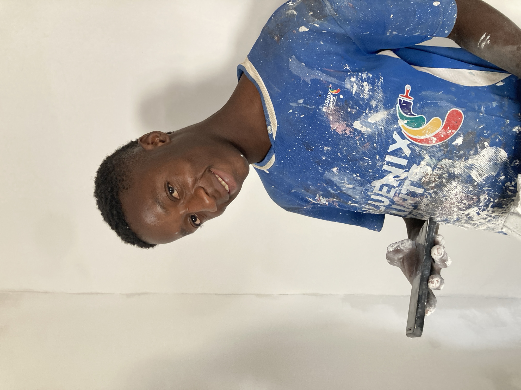A man is holding a rectangular object, likely a smartphone. He is wearing a blue shirt with white paint splatters. The shirt has the logo "BLUENIX PAINTS" on it. The background is a plain white wall, possibly freshly painted. The location may be Bukuru, Nigeria, based on information related to Bluenix Paints' presence in the area.
