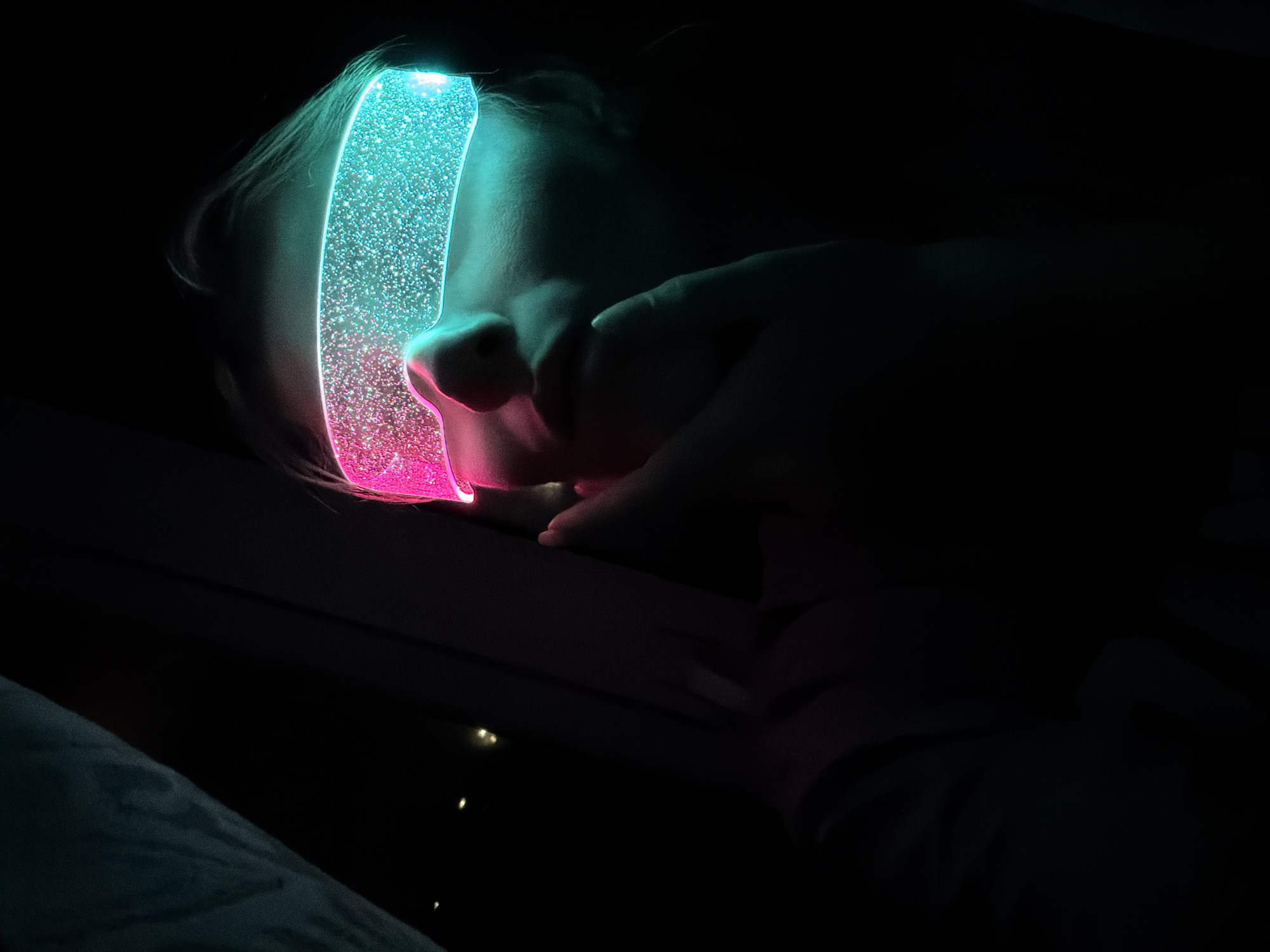 A low-light, close-up photograph depicts a person, likely a child, seen in profile, resting their head against a dark surface. The focal point is a vibrant, transparent, and glitter-filled object worn across the person's forehead. This head-mounted device emits a gradient of light, transitioning from a bright aqua or teal color at the top to a vivid magenta or pink at the bottom. Tiny, reflective particles within the object sparkle intensely.
The glowing device illuminates parts of the person's face, including their forehead, bridge of the nose, and a portion of their cheek, casting colorful hues onto their skin and light-colored hair. A hand, presumably the subject's, is visible in the lower right quadrant, resting gently on a dark surface. The background is almost entirely black, indicating a very dark indoor environment, such as a bedroom at night. Faint, small points of light can be seen in the deeper background. A hint of bluish-grey fabric, possibly a blanket, is visible in the bottom left corner. The scene evokes a sense of quiet wonder and peaceful rest, with the luminous object providing the sole significant light source. No text is visible in the image. The setting is an indoor space in Chaikovsky, Russia.