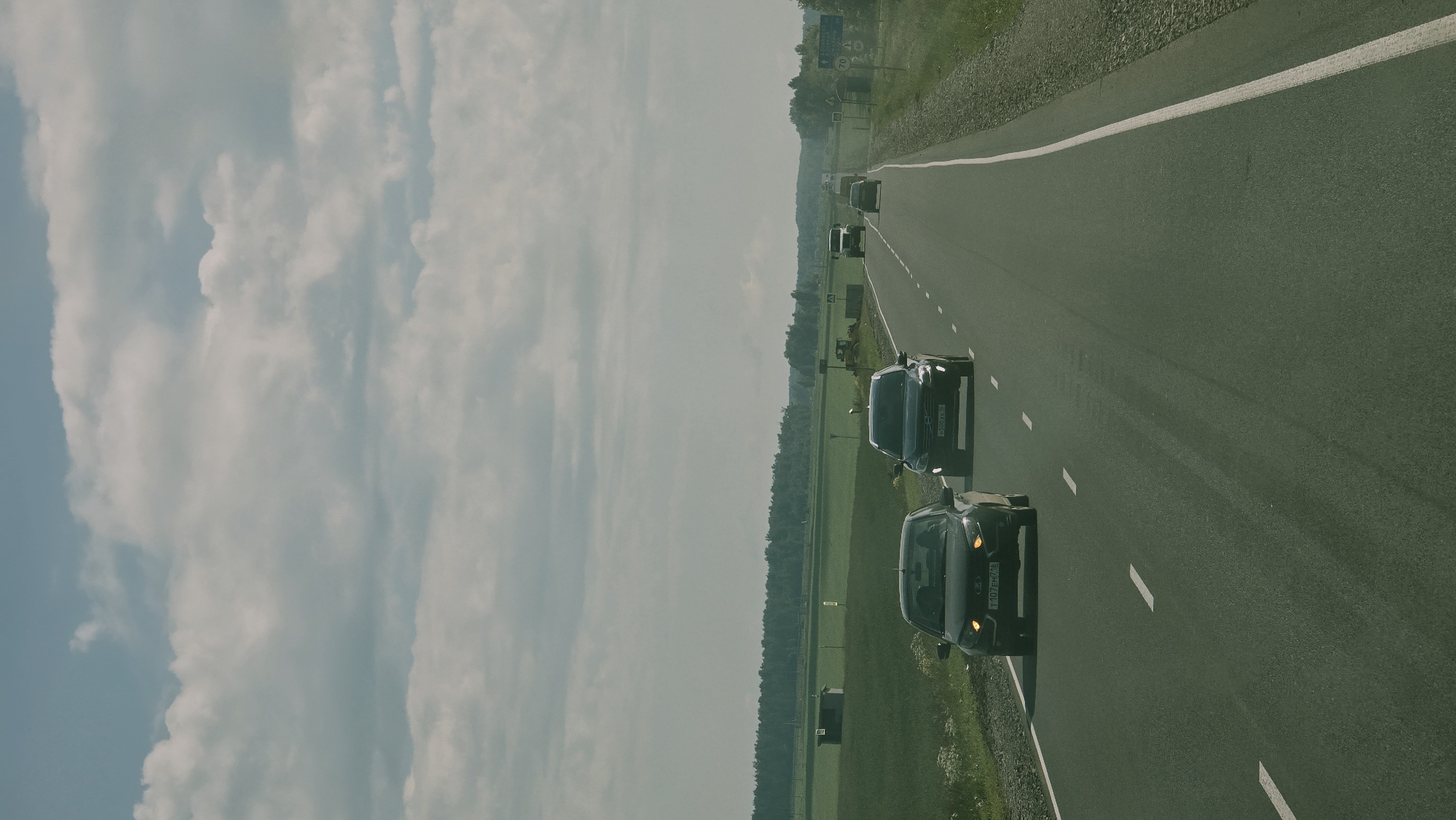 A daylight scene on a rural highway shows several cars driving in the same direction. In the foreground, a dark-colored sedan with license plate T107EM716 is on the left lane. To its right and slightly ahead, a dark SUV with license plate A500XX86 is in the right lane. Further ahead on the right lane, a white truck is visible. The highway is paved and has white dashed lines dividing the lanes. The surrounding environment consists of a grassy field on the left with a small wooden structure and a treeline in the distance. On the right, there is a gravel shoulder and more vegetation. The sky is partly cloudy with large, white clouds. The scene appears to be in a rural area of Актюбинский, Russia, based on the license plates.