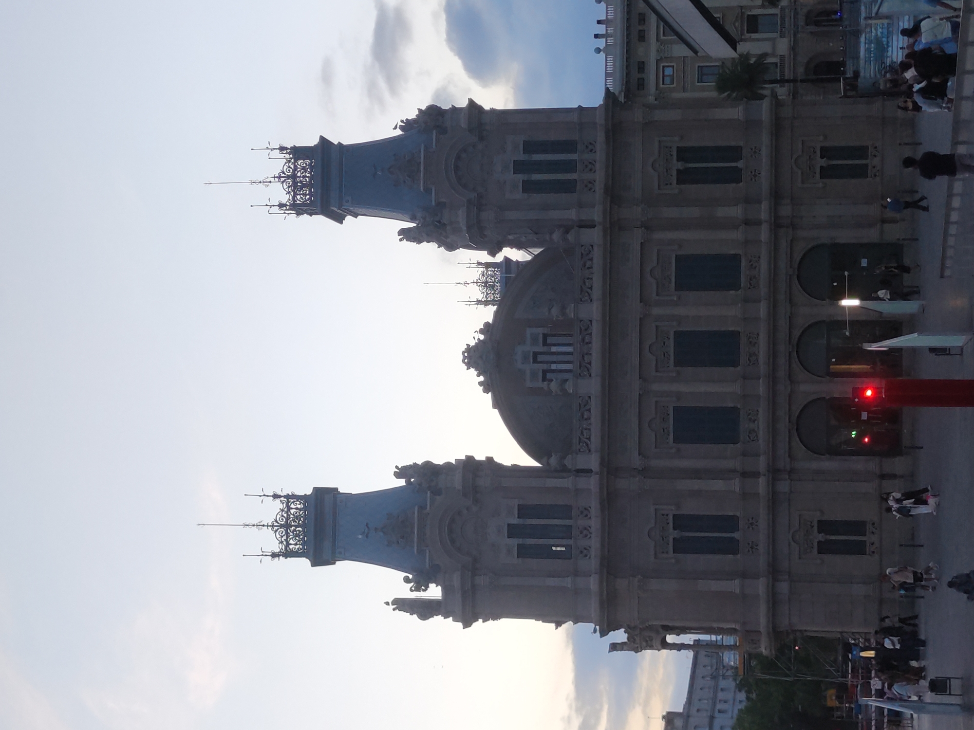 This image depicts the exterior of a large, ornate building with two prominent towers, likely a public building or a historical landmark. The architecture suggests a classical or neoclassical style, characterized by intricate detailing, columns, arched windows, and decorative spires atop the towers. The building is constructed from stone, with a muted, possibly gray or beige, facade.
The scene takes place outdoors, likely in a public square or plaza, as evidenced by the paved ground, a red traffic signal, and several individuals walking or standing around. The time of day appears to be dusk or early evening, as the sky is a pale, hazy blue with some clouds, and the lighting is soft.
In the foreground, a paved area is visible with several people scattered throughout. Some are walking in groups, while others appear to be individuals observing the surroundings. A red traffic light is prominently displayed in the middle of the scene, suggesting proximity to a road or intersection.
In the background, the grand building dominates the frame. Adjacent to it, to the right, a modern structure with a slanted roofline is partially visible, indicating a contrast between historical and contemporary architecture. Further in the background, more buildings can be seen, suggesting an urban environment.
The image does not provide specific visual cues to pinpoint the location to Sant Boi de Llobregat, Spain. However, the architectural style and the general urban setting are consistent with many European cities.