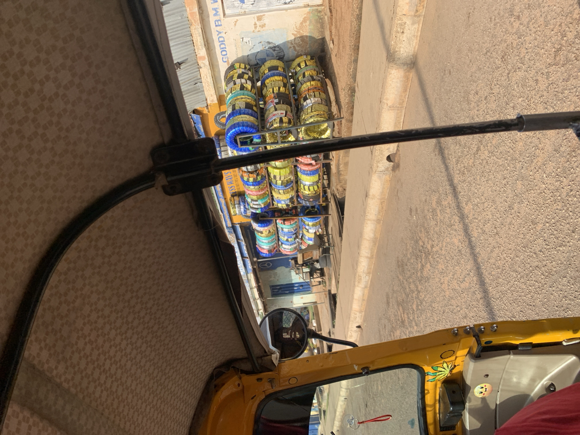 The image appears to have been taken from inside a yellow auto rickshaw, possibly near New Stadium in Nigeria, judging by the architectural style of the adjacent building. The view is from the driver's seat, looking out towards the street. Stacks of tires, wrapped in various colored plastic, are displayed for sale outside a storefront labeled "GODDY B.M.W." The street is paved. The interior of the auto rickshaw is visible, including the checkered pattern of the ceiling and the side mirror. A portion of the rickshaw's frame is also visible in the foreground. The overall perspective is slightly tilted, suggesting the photograph may have been taken on the move or with a mobile device.