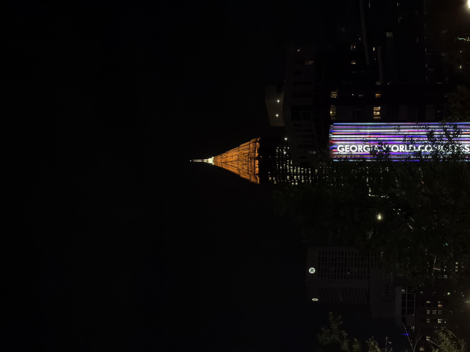 The image depicts a night scene in Atlanta, United States of America. The Georgia World Congress Center is identifiable by its illuminated signage. In the background, a tower with its spire is visible in yellow lights. Trees are in the foreground. The image is oriented upside down.