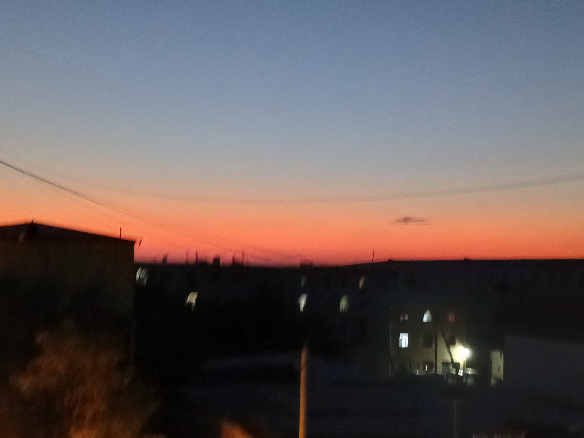 A dusk scene captures the sky over Qorowul, Uzbekistan, transitioning from a soft blue to a vibrant orange gradient. Silhouetted against the colorful sky are the dark forms of buildings, some with illuminated windows. A faint wire traverses the sky, and a small, indistinct cloud is visible in the upper right quadrant. The foreground is dominated by the dark shapes of structures, with hints of foliage in the lower left. The overall impression is one of a quiet urban evening.