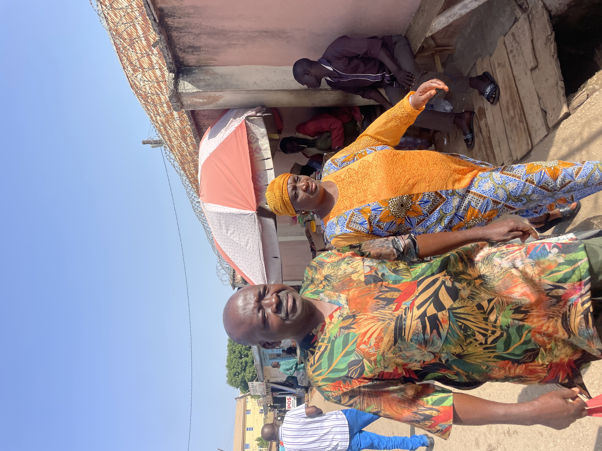 The image captures a street scene in Gura Top, Nigeria, featuring two individuals in the foreground. A man in a colorful, tropical-patterned shirt stands alongside a woman adorned in a vibrant orange top and patterned blue pants, with a matching head wrap. Behind them, several people are seated under the partial shade of a building. An open, striped umbrella provides additional shading. A building with a corrugated roof and barbed wire is visible in the upper left. In the background, a street scene with buildings and pedestrians is discernible under a clear blue sky.