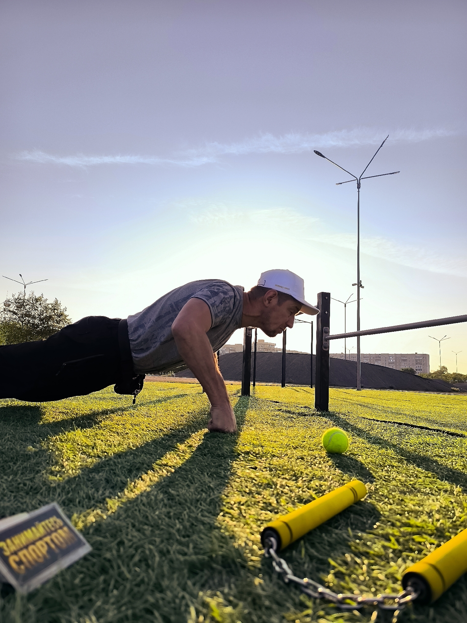 A man is performing a push-up on an outdoor sports field with artificial turf. He is wearing a grey t-shirt, black pants, and a white baseball cap. A yellow tennis ball and a pair of yellow nunchaku are lying on the grass nearby. In the background, there are streetlights, buildings, and a clear sky. The scene appears to be a public park or sports complex, possibly located within the city administration area of Qarağandı, Kazakhstan, given the presence of urban infrastructure and the distinctive architecture of the buildings in the distance. The lighting suggests it is either early morning or late afternoon, with the sun casting long shadows across the field. A small sign with yellow text on a dark background is visible in the foreground, which reads "ЗАНИМАЙТЕСЬ СПОРТОМ" (Engage in Sports).