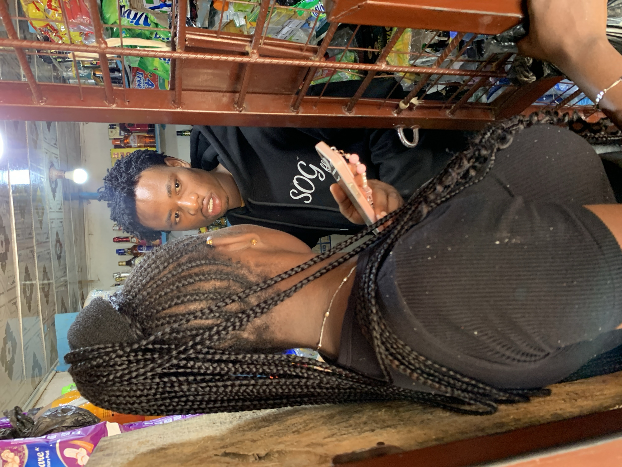 The image shows two young individuals in what appears to be a small shop or market stall, possibly located in Jos, Nigeria. The individual in the foreground, viewed from the back, has long, dark braided hair and is wearing a black top. They are holding a light pink smartphone with a decorative keychain. Facing this individual is a young man with short, curly dark hair, wearing a black hoodie with the white text "SOG" across the chest. He is looking intently at the phone being held by the other person.
The environment is cluttered with various goods displayed on shelves behind a metal grate. These items include bottles, bags of snacks or food products, and other packaged goods, suggesting a retail setting. The lighting is somewhat dim, with a bright overhead bulb visible. The overall impression is of a candid moment captured within a busy, local marketplace.