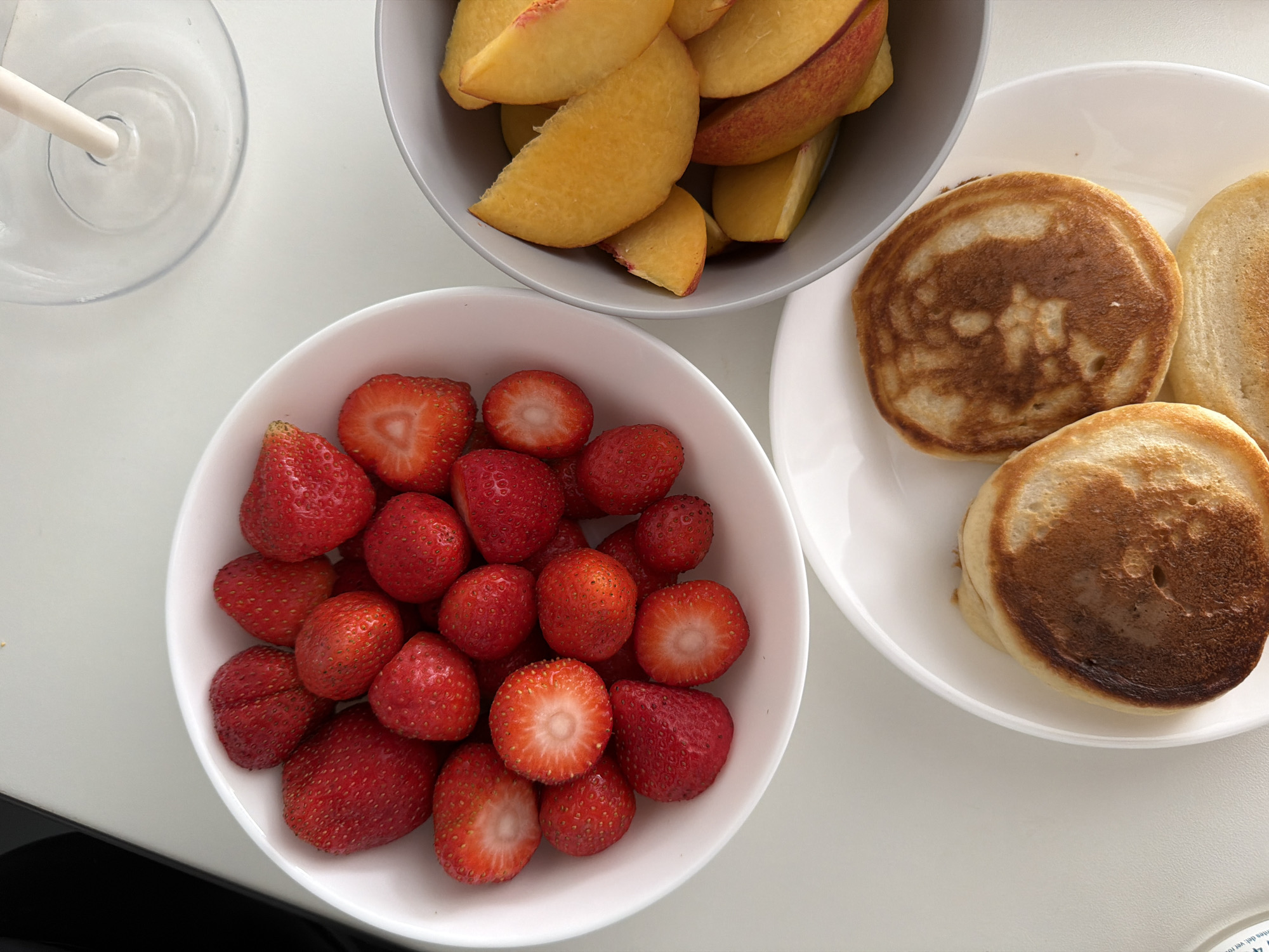 A top-down, close-up shot shows a breakfast spread on a white surface. In the center left, a white bowl is filled with fresh, red strawberries. To the right, a grey bowl contains sliced peaches with yellow flesh and reddish skin. Below these bowls, on a white plate, are three small, golden-brown pancakes. The scene is well-lit, highlighting the textures and colors of the food. The setting appears to be a domestic environment, possibly a kitchen or dining area, in Finestrat, Spain. No people are visible in the image.