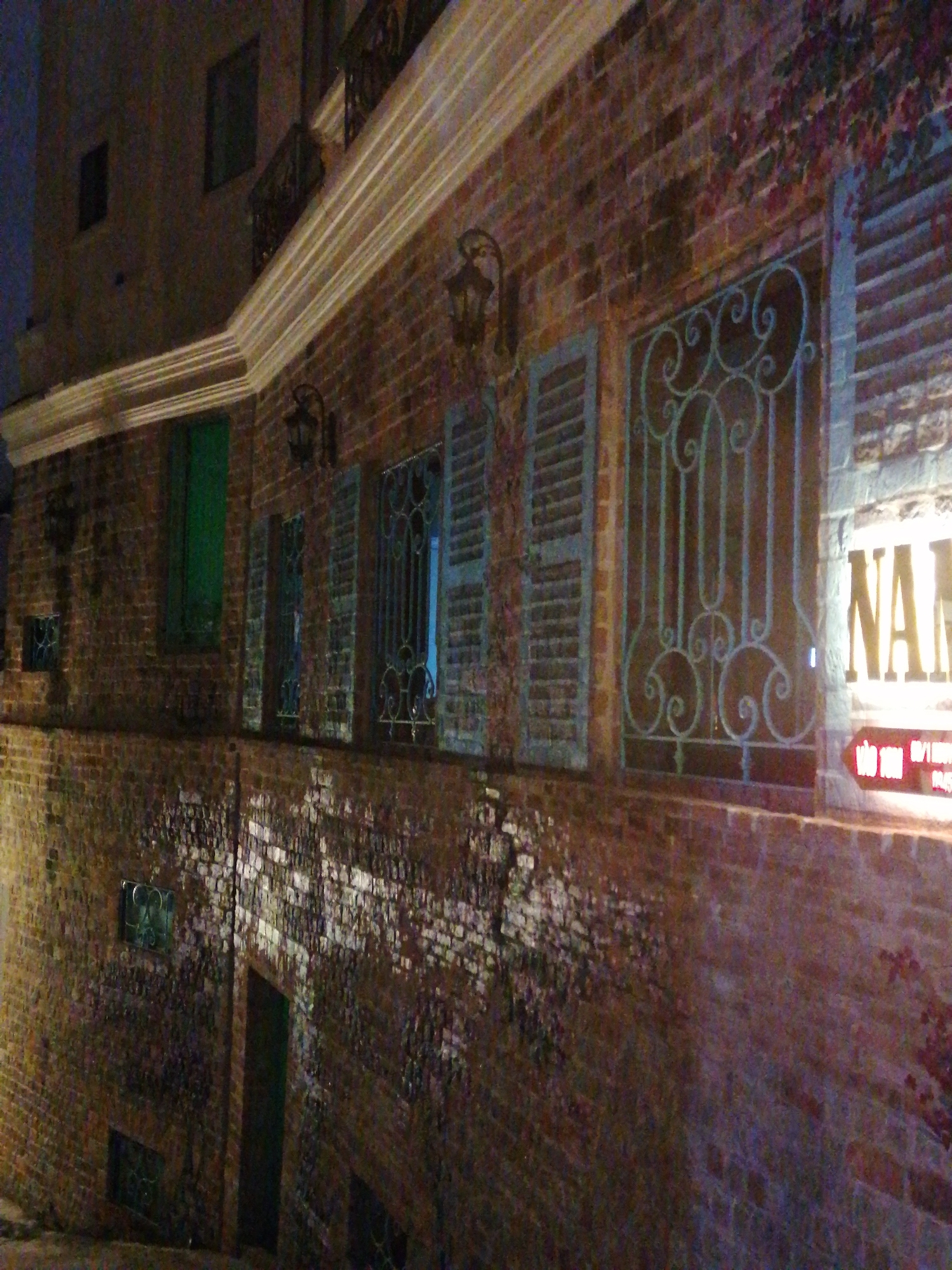 The image captures the facade of a brick building at night, illuminated by artificial lights. The building features a classic architectural style, with prominent red-brown brickwork. A long, light-colored cornice or molding runs horizontally across the building's upper section, catching the light.
Numerous windows are visible, many equipped with light-colored, louvered shutters that are either closed or partially open. Several windows are adorned with intricate, dark-colored wrought iron grilles featuring decorative scrolls and patterns. On the upper left, glimpses of windows and balconies with ornate railings can be seen. Wall sconces or lanterns are mounted on the brick walls, adding to the period aesthetic.
A large, illuminated sign on the right side of the building displays the partial text "NA" in bright, light-colored letters, likely part of a larger name. Below this, a red arrow points left, and there is smaller text in Vietnamese, which appears to include "VÀO HÀNG" and numbers like "0911 30..." and possibly "0984".
Some sections of the brick wall, particularly lower down, show lighter patches, possibly from reflected light or dampness, and hints of climbing vines or foliage are visible near the top right window. A dark green window or door is noticeable on the left side of the building, and another dark door is seen at ground level on the lower-left side. The overall scene suggests a commercial establishment or residence, possibly in a historic district, consistent with a setting in Da Lat, Vietnam.