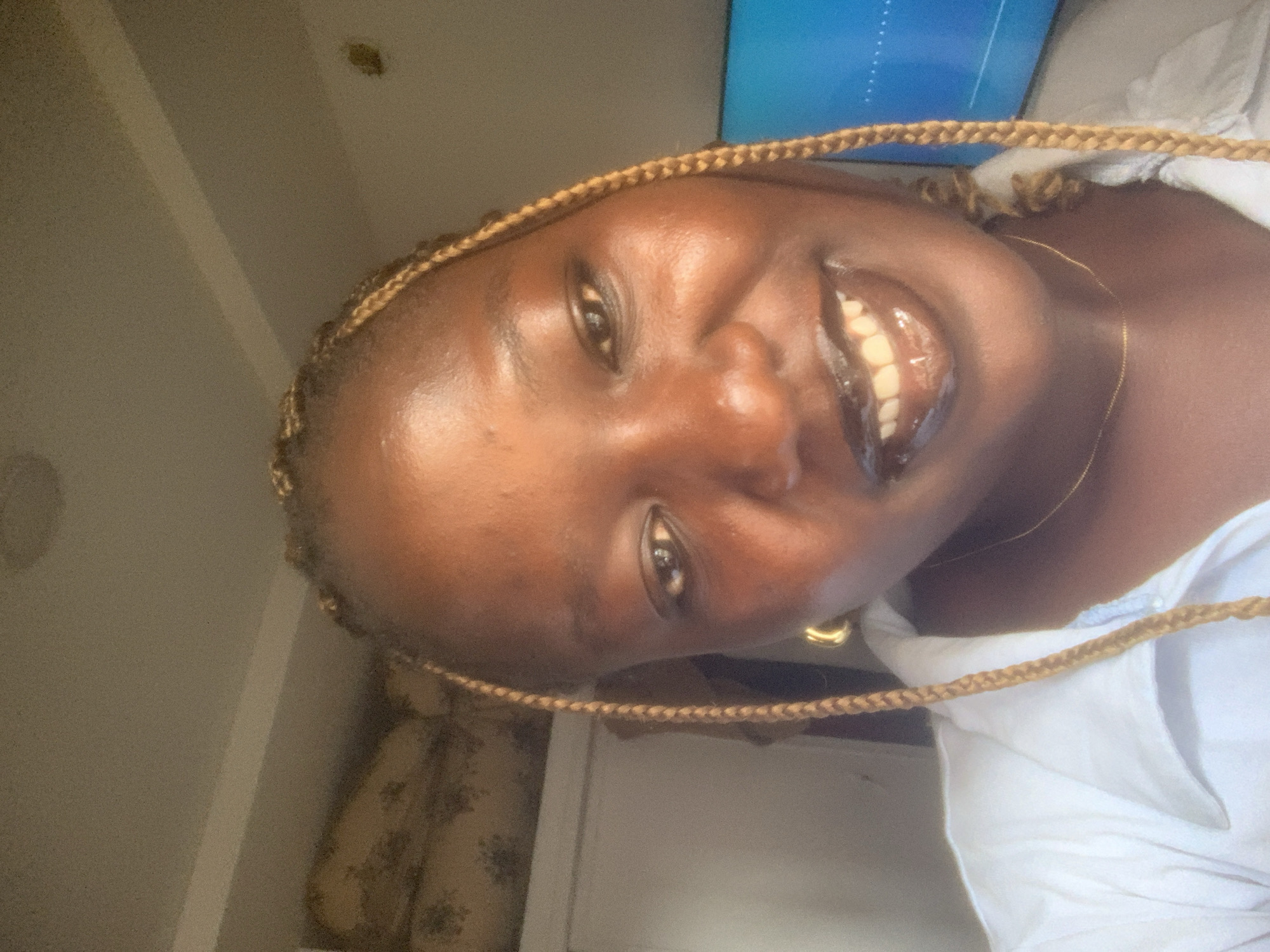 A close-up, slightly tilted portrait shows a dark-skinned woman with blonde braided hair framing her face. She is smiling, exposing her teeth. She is wearing a white top and a gold chain necklace. A gold hoop earring is visible in her left ear. Behind her, a beige wall is partially visible, along with a beige patterned cushion. A television screen emitting a blue glow can be seen in the upper right corner. The exact location within Ado, Nigeria, cannot be determined from the image.