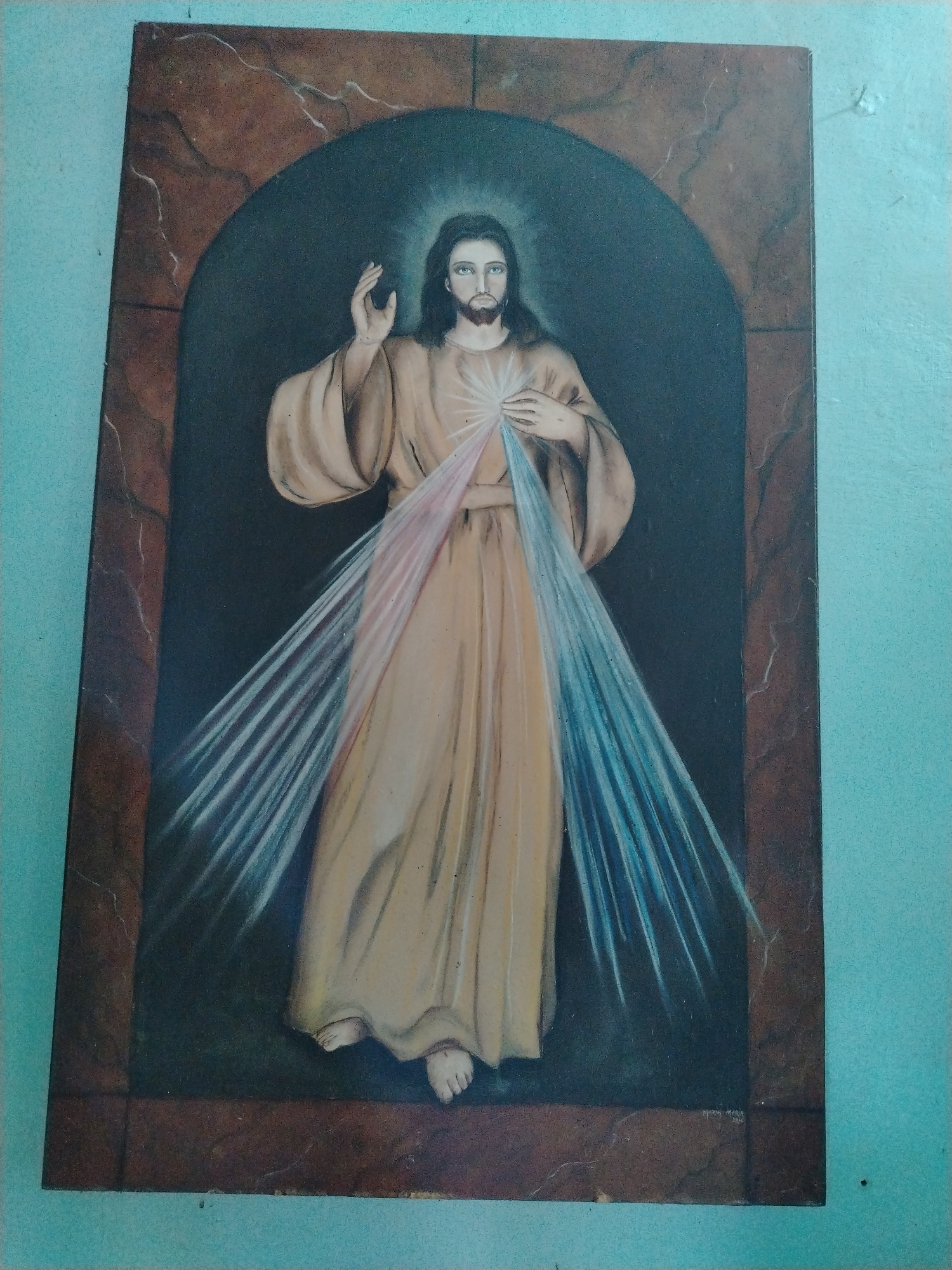 The image depicts a painting of Jesus Christ, likely the Divine Mercy image. He is shown standing, wearing a long, flowing robe with pink and blue rays emanating from his heart area. His right hand is raised in a blessing gesture, and his left hand is touching his chest. The painting is framed, with the frame having a faux marble or stone texture. The painting is hanging on a wall painted in light blue. The location is Los Leoncitos, Venezuela, based on the presence of a Maria Maria 2010 signature on the bottom of the painting. There is no other visible setting information.