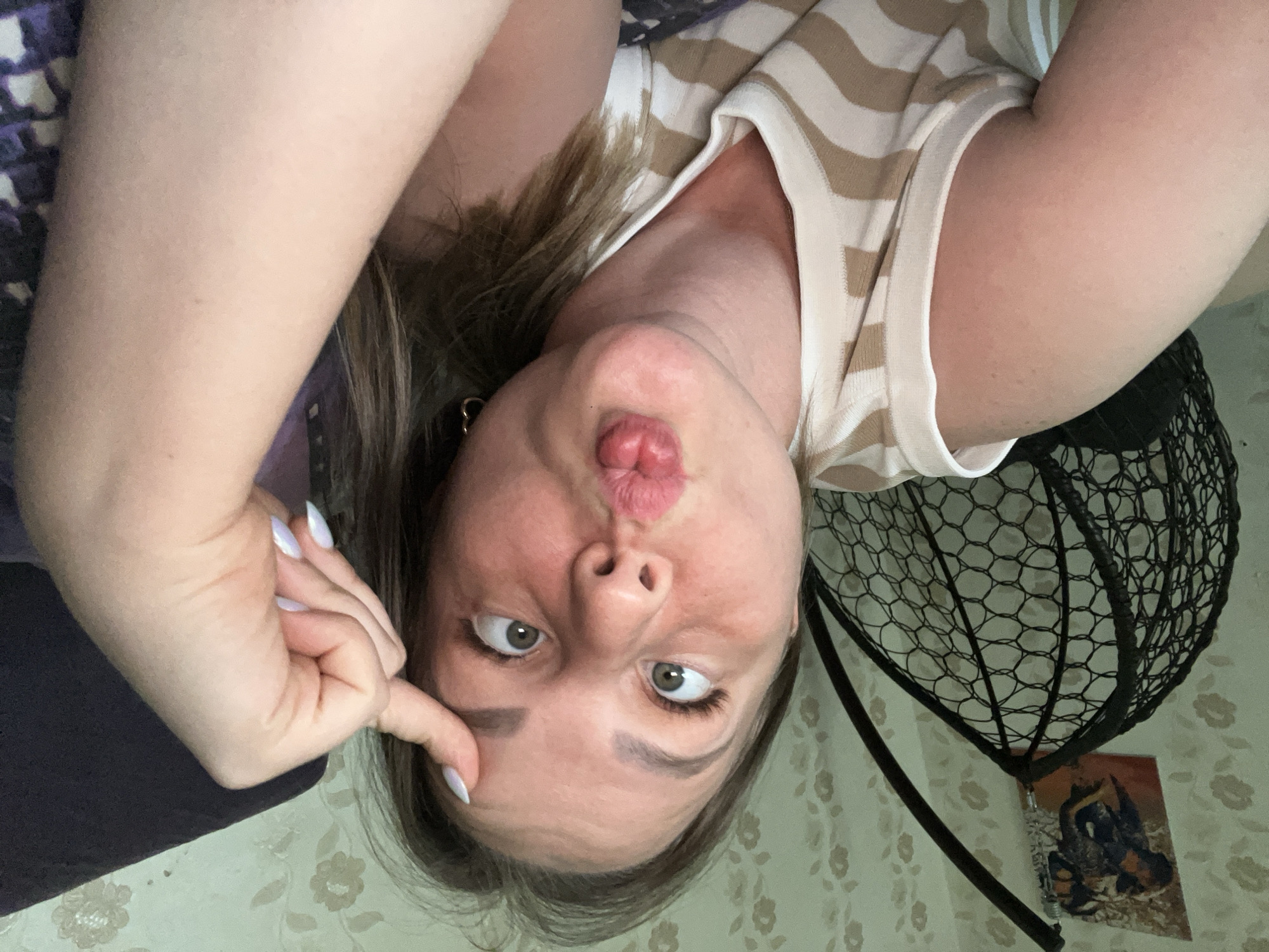 A woman is holding her finger to her forehead and making a kissing face at the camera. She is wearing a white shirt with light brown stripes. Behind her is a patterned wall and a black wire basket. There is no information to indicate this image was taken in Izhevsk, Russia.