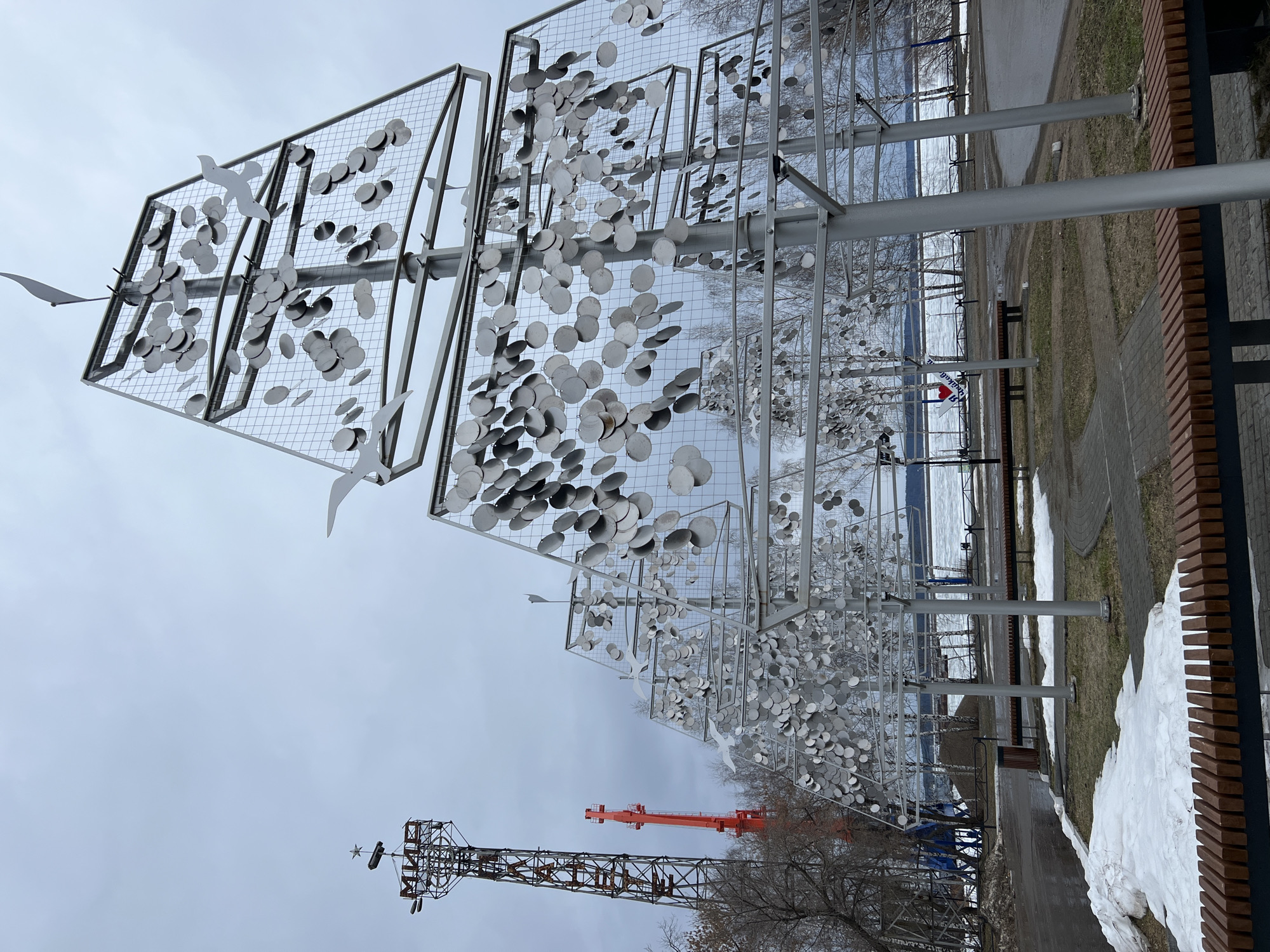 The image depicts a public art installation composed of multiple square frames filled with metal discs and bird cutouts, arranged in a row along a pedestrian walkway. The frames are supported by metal poles. In the background, a body of water is partially visible, with snow on the ground indicating winter or early spring. In the distance, an industrial tower is noticeable. The sky is overcast. This art piece is located in the city of Chaikovsky, Russia. The immediate surroundings consist of a paved walkway, a grassy area, a low brick wall, and remnants of snow. There are no people visible in the image.