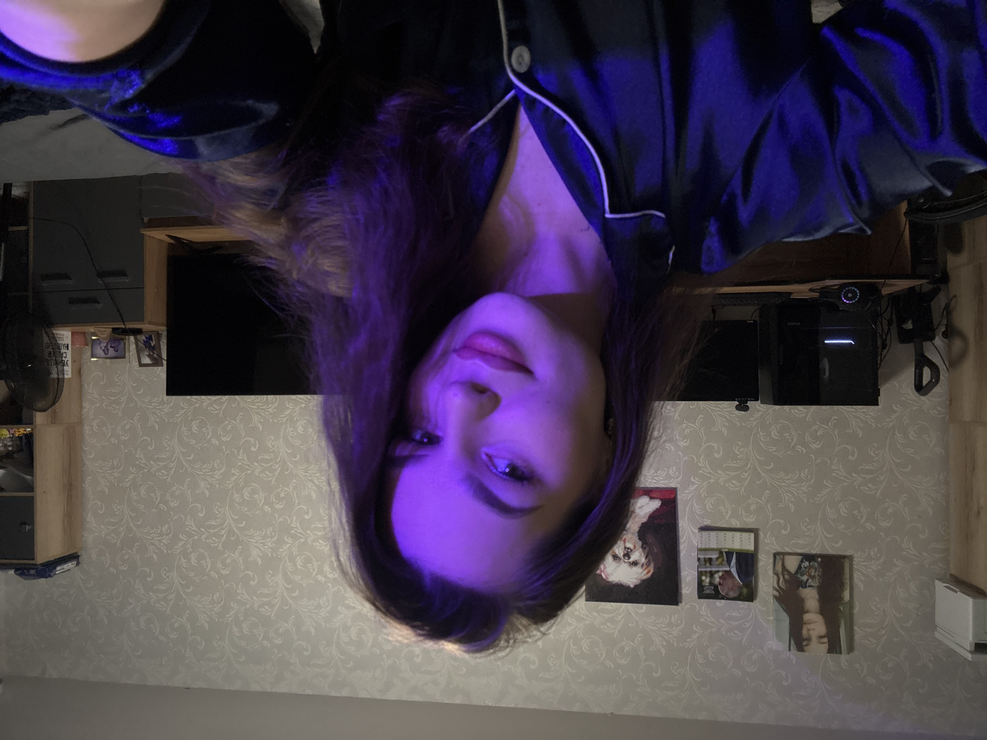A medium shot captures a young woman with long, dark hair, illuminated by purple lighting, as she looks directly at the camera. She is wearing dark pajamas with white trim. The background shows a bedroom with patterned wallpaper. On the wall, there are framed pictures of a woman, a calendar with a man's image, and a painting of a dog. To the left, a closet and a computer setup are visible. To the right, a television and a dresser are present. The overall setting appears to be a domestic environment in Chaikovsky, Russia.