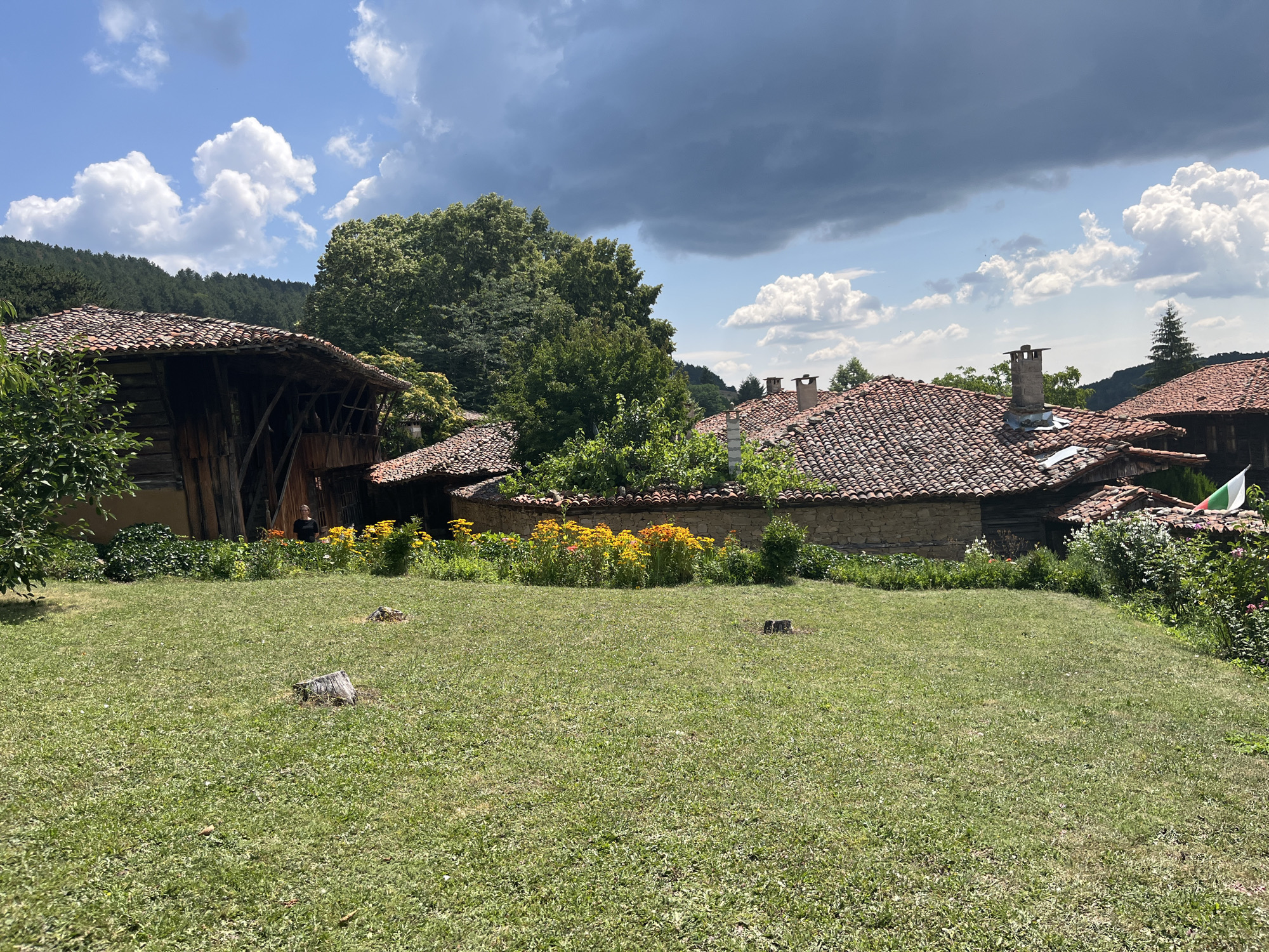The image shows a scene in the village of Zheravna, Bulgaria. In the foreground, a well-maintained green lawn with several tree stumps is visible. In the middle ground, a low stone wall separates the lawn from a garden area featuring a vibrant bed of yellow and orange flowers. Behind the flowers, traditional Bulgarian houses with red tiled roofs and stone walls are seen. One house on the left has a distinctive wooden structure and a dark opening. A person is standing near the opening of this house. Other houses are visible further back, with some featuring chimneys and the Bulgarian flag. The background consists of lush green trees and a hillside, under a partly cloudy sky with dramatic dark clouds.
