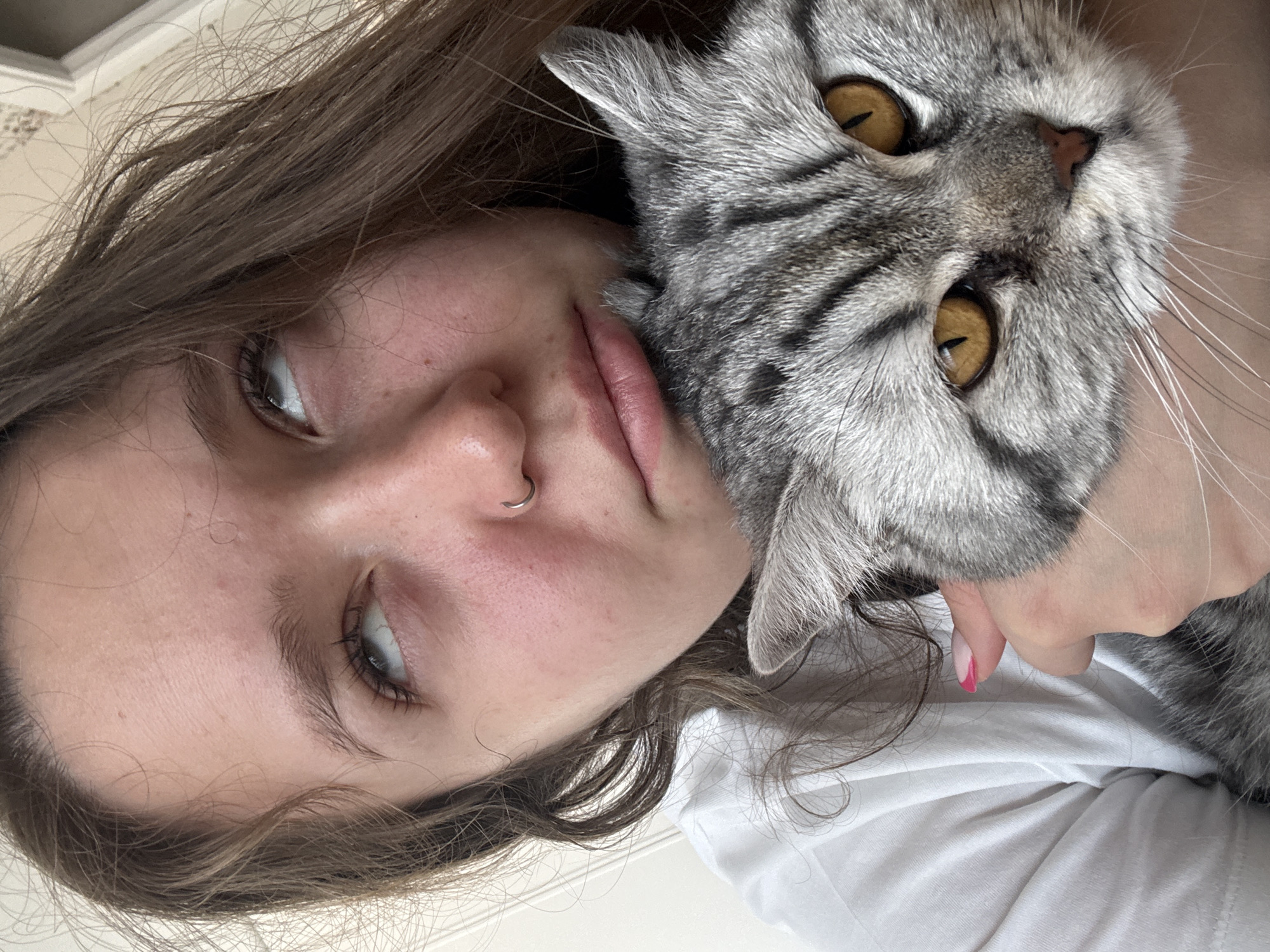 A close-up, tilted shot shows a young woman holding a gray tabby cat close to her face. The woman has light brown hair and light skin. She has a silver nose ring in her left nostril. Her lips are slightly parted. The cat has yellow eyes and gray and black striped fur. The woman is wearing a white shirt. The background is slightly blurred and appears to be a wall with a subtle pattern. There is no information to locate this image in Izhevsk, Russia.