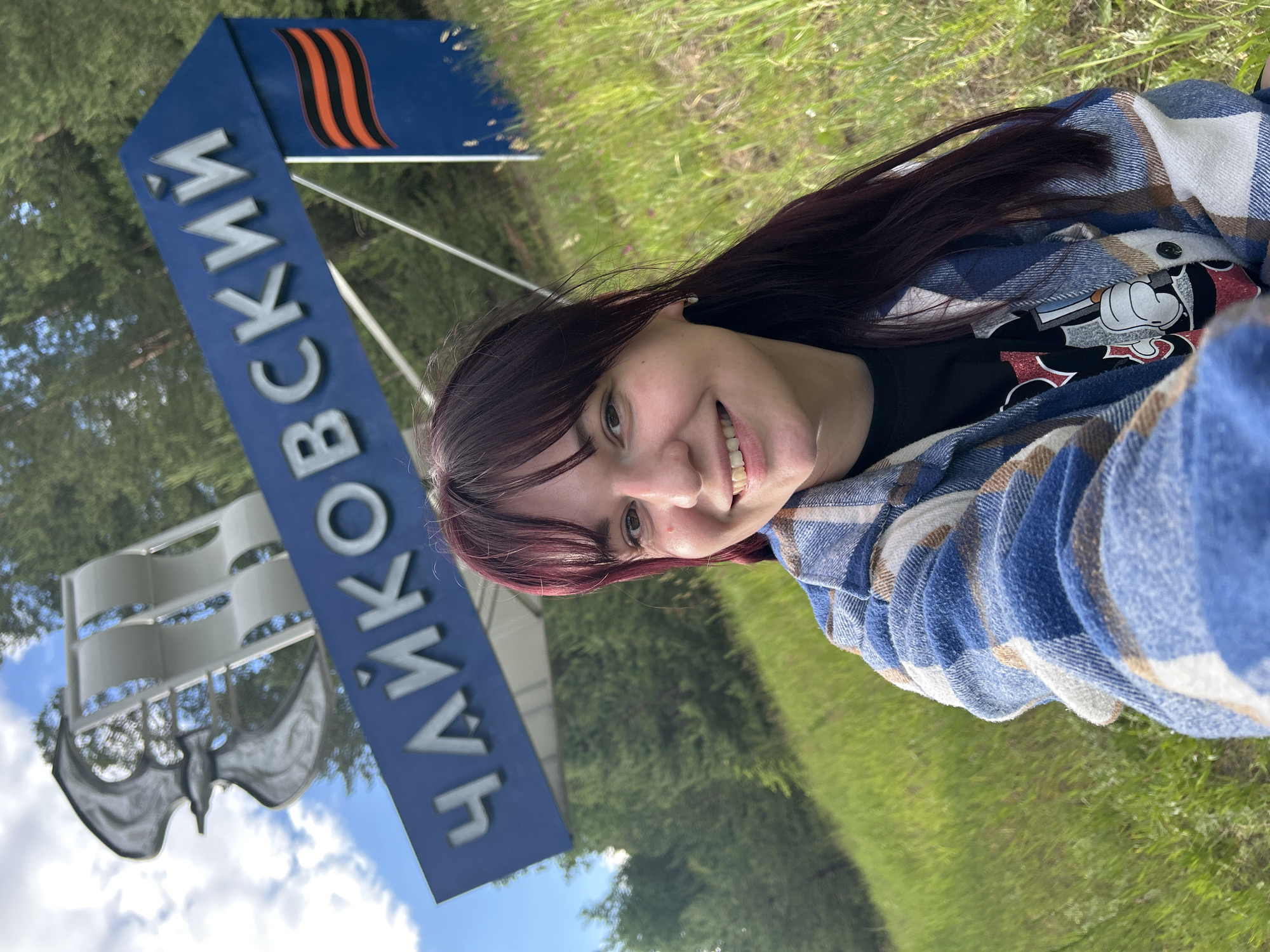 A young woman with long, dark, purple-tinted hair and bangs is taking a selfie outdoors. She is wearing a blue and white plaid shirt over a black t-shirt with a graphic of Mickey Mouse. She is smiling and looking at the camera. Behind her, a large blue sign reads "ЧАЙКОВСКИЙ" in silver letters, indicating the city of Chaikovsky, Russia. To the right of the sign, there is a vertical blue panel with an orange and black striped ribbon, possibly representing the Order of St. George. A silver metallic sculpture resembling wings or sails is also visible above the sign. The background consists of a green, grassy hillside with scattered trees and a blue sky with white clouds.
