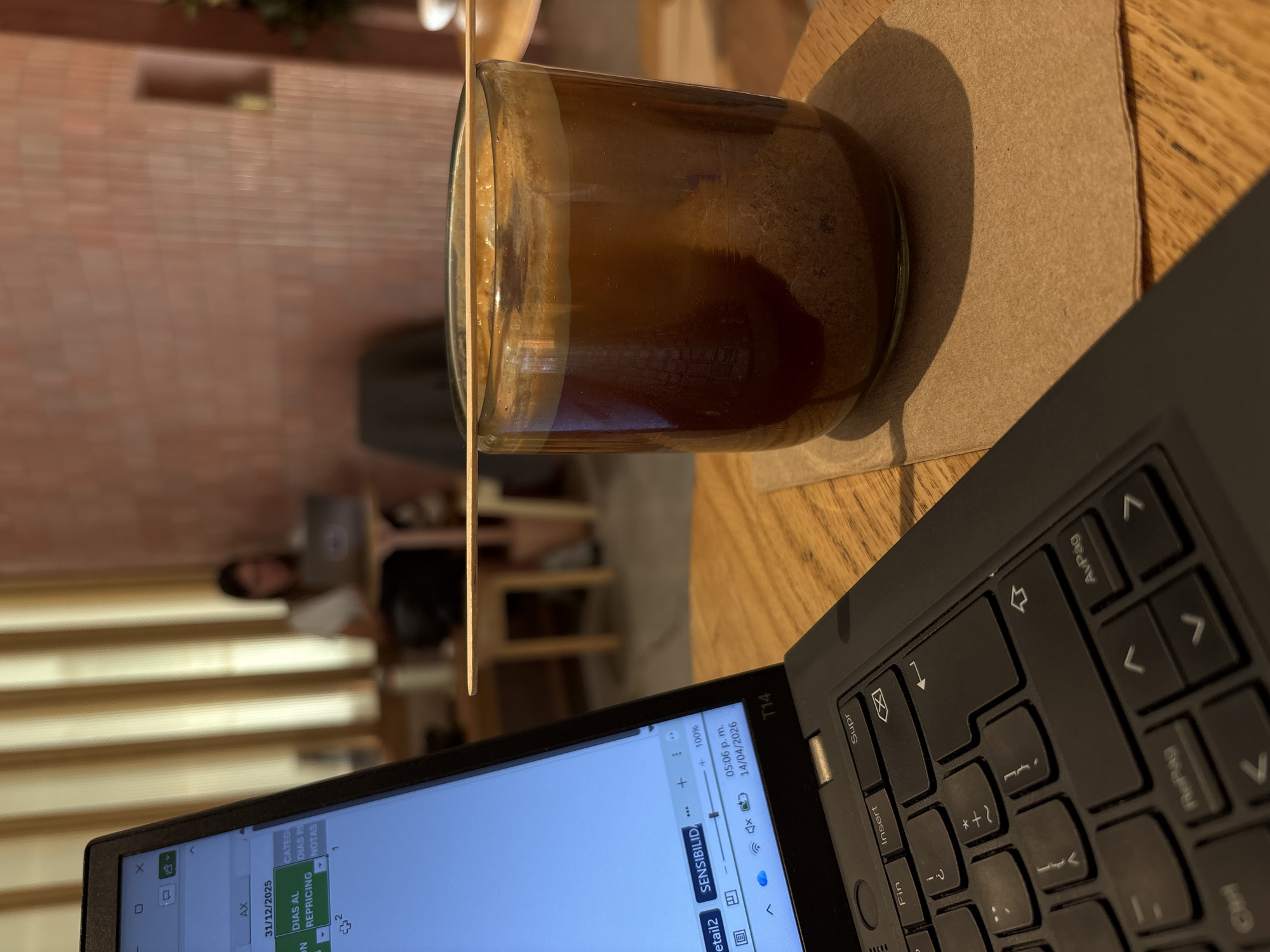 The image captures an indoor setting, likely a cafe or workspace. A laptop, partially visible in the lower right corner, displays a spreadsheet, indicating work or study activity. Adjacent to the laptop is a glass filled with a dark liquid, possibly coffee, placed on a brown coaster. A wooden stirrer protrudes from the drink. In the background, a person is seated at another table, working on a laptop, suggesting a shared workspace or public cafe environment. The foreground table and background settings imply a relaxed yet productive atmosphere.