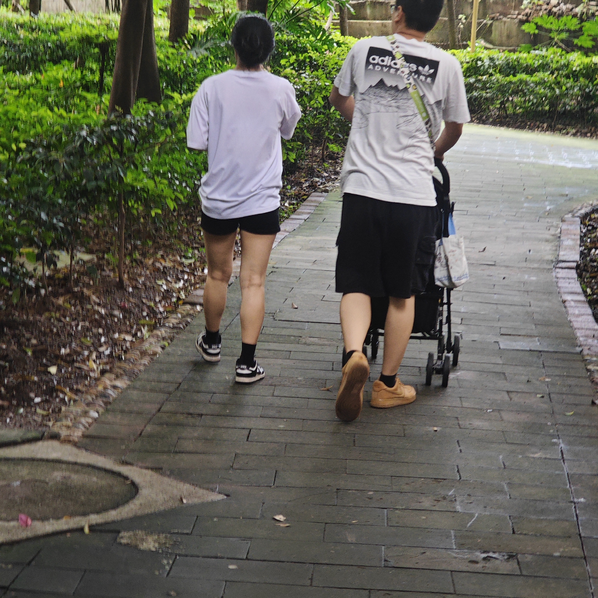 A photo taken at ground level shows two people walking away from the camera on a brick pathway, pushing a stroller. The location is outdoors in Yuzhong District, China.
The person on the left wears a white t-shirt, black shorts, black socks, and black and white sneakers. The person on the right wears a white t-shirt with an Adidas logo on the back, black shorts, black socks, and tan sneakers. They push a black stroller with a bag attached. The path is made of dark gray bricks and is surrounded by foliage. There are trees and bushes visible. There is a circle of dirt in the bottom left corner.