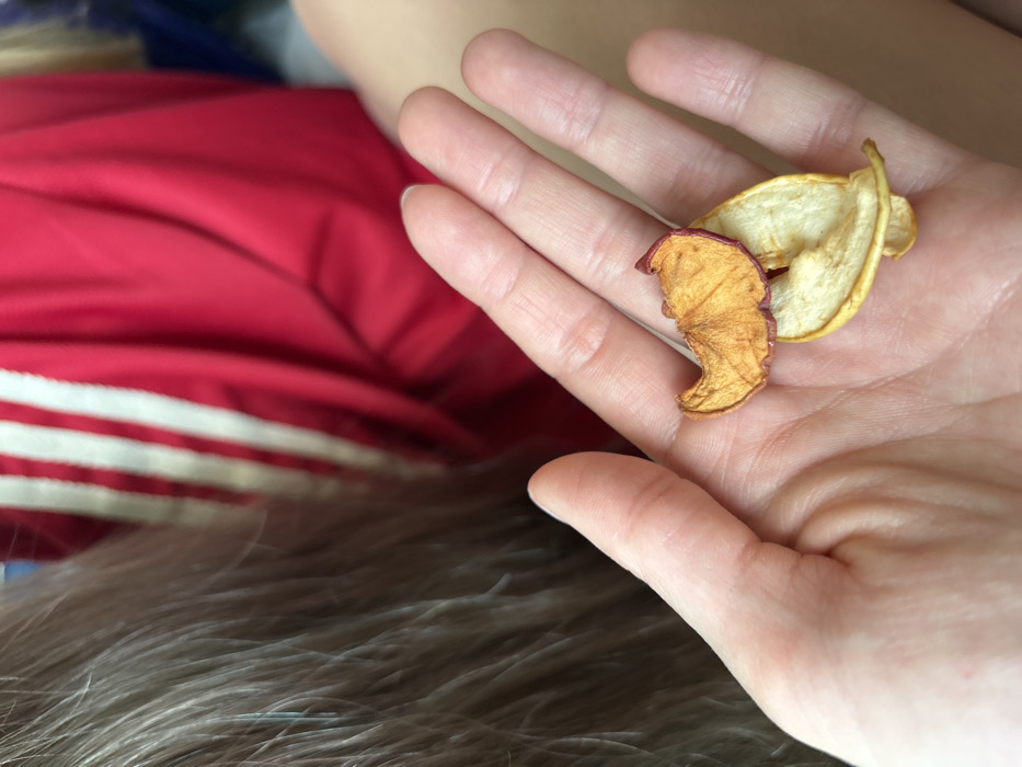 The image captures a close-up, downward-angled view of a human hand presenting three dried apple slices against a blurred background. The hand, positioned in the lower-center, has its palm open and upturned, holding two pale yellow-cream dried apple slices overlaid by a third, darker reddish-orange slice with a visible red peel edge. In the upper left background, out of focus, is a mass of light-colored hair. To the right and behind the hand, also out of focus, is a segment of a red garment featuring two prominent vertical white stripes, consistent with athletic wear, and an expanse of light-toned bare skin, possibly a leg or thigh. The scene is located in Ivanovka, Russia.