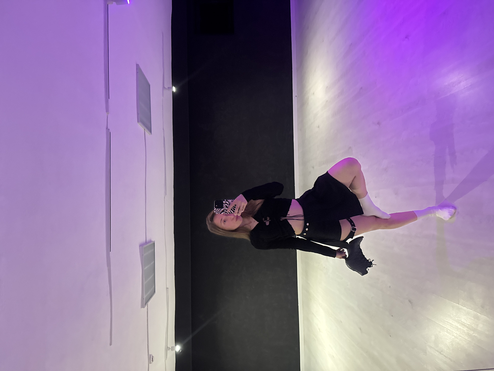 A young woman is captured in a full-body selfie taken in an indoor setting, likely a studio or modern space. She has long, straight blonde hair and is looking directly at the camera with a neutral expression, holding a smartphone with a distinctive zebra-print case in her right hand.
She is dressed in a contemporary, somewhat edgy outfit consisting of a black long-sleeved top with cut-out details and ties at the front, paired with a black mini-skirt. A black garter belt or leg harness is visible on her upper right thigh. Her footwear is mismatched: she wears a white sock or boot on her left leg and a black athletic-style shoe on her right leg.
The background is stark and modern, divided into distinct sections. To her left, there is a white wall featuring several rectangular recessed panels or vents. Behind her is a solid black wall, while the area to her right and beneath her feet is a light-colored floor or wall surface. This area is dramatically illuminated with purple and white lighting, casting a faint shadow of a person onto the surface. There are also small, bright light fixtures visible on the walls. The overall ambiance is artistic and urban, created by the contrasting colors and specialized lighting. No visible text is present in the image.