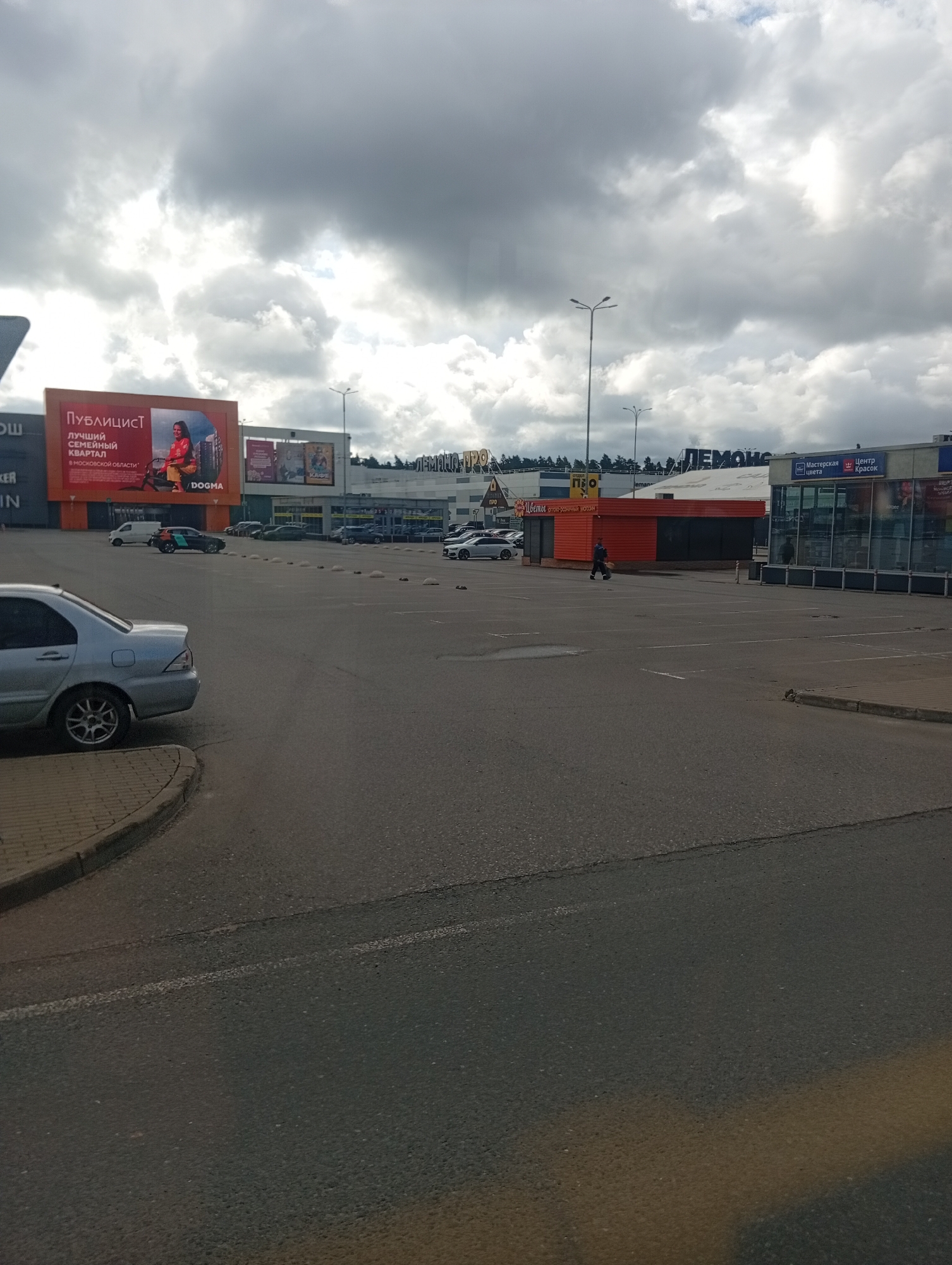 The image shows a street view in Pushkino, Russia. In the foreground, a silver sedan is parked on the left side of a paved parking lot. The lot is mostly empty, with a few other cars visible in the distance. In the background, a large shopping center complex is visible, with various storefronts and illuminated signs. One sign prominently displays "ПУБЛИЦИСТ" (Publicist) with an image of a woman and text in Russian. Another large sign reads "ЛЕМОН" (Lemon). The sky is overcast with dramatic, dark clouds. A person is walking in the middle distance across the parking lot. The overall scene depicts a commercial area under a cloudy sky.