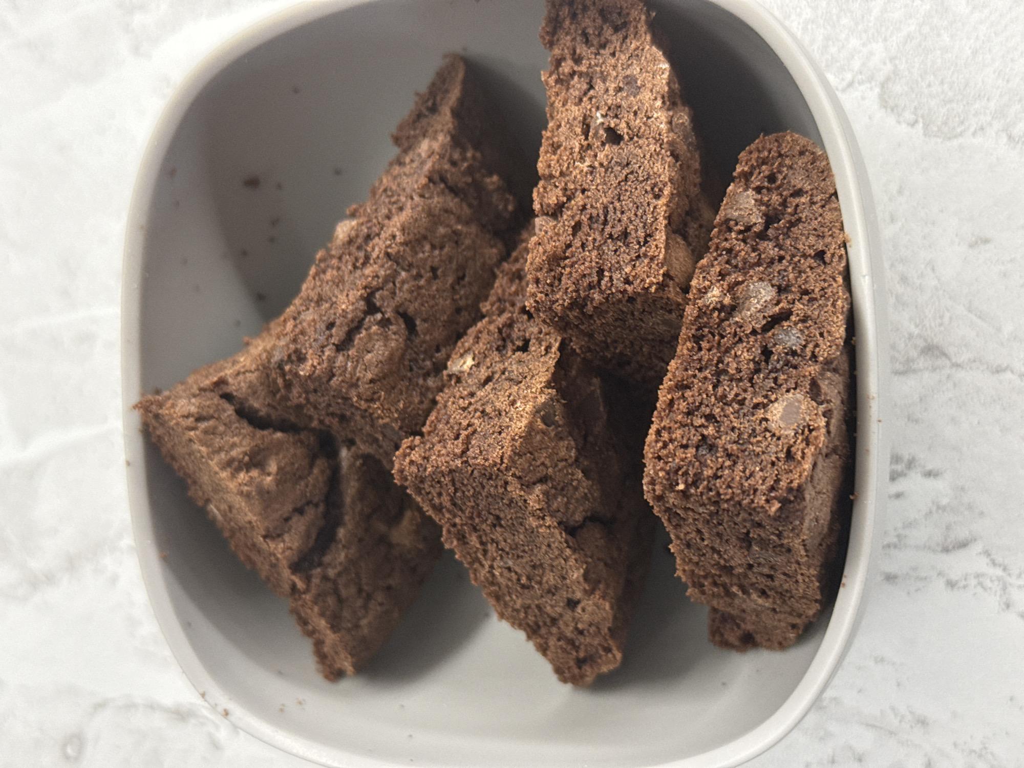 A close-up, overhead shot shows several chocolate biscotti pieces in a light gray, shallow bowl. The biscotti are triangular and appear to be a deep brown color with visible chocolate chips embedded within. The bowl is placed on a textured white surface, possibly a marble countertop. The lighting casts subtle shadows, highlighting the crumbly texture of the biscotti. There are no people or animals present in the image, and no discernible location within the city of Tomilino, Russia can be identified.