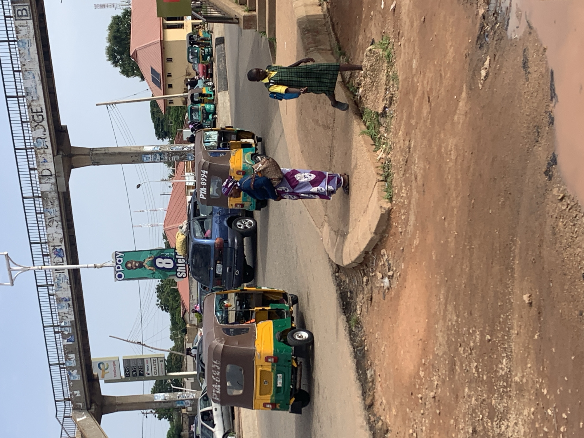 The image depicts a street scene in Gura Top, Nigeria, under a bright, possibly overcast sky. A pedestrian bridge, weathered and marked with graffiti, spans the roadway. Below the bridge, a gas station sign for TotalEnergies is visible. In the foreground, a woman wearing a headscarf and a colorful, patterned wrap dress carries a large bag over her shoulder. She stands on a raised concrete walkway. To her right, a young girl in a green plaid school uniform with a blue backpack walks along the same walkway, looking towards the right of the frame. Several auto-rickshaws, commonly known as "Keke Napeps" in Nigeria, are present on the road. One is yellow and green, with the license plate PTA-6636. Another has PTA-8994 on its rear. A blue SUV is parked on the road behind the Keke Napeps, and further down the road, several other Keke Napeps and a red car are visible. Buildings with red-tiled roofs line the street in the background, with trees and utility poles also present. The ground in the immediate foreground appears to be unpaved, reddish-brown earth with scattered debris and a puddle of muddy water. The overall impression is of a busy, functional urban environment.