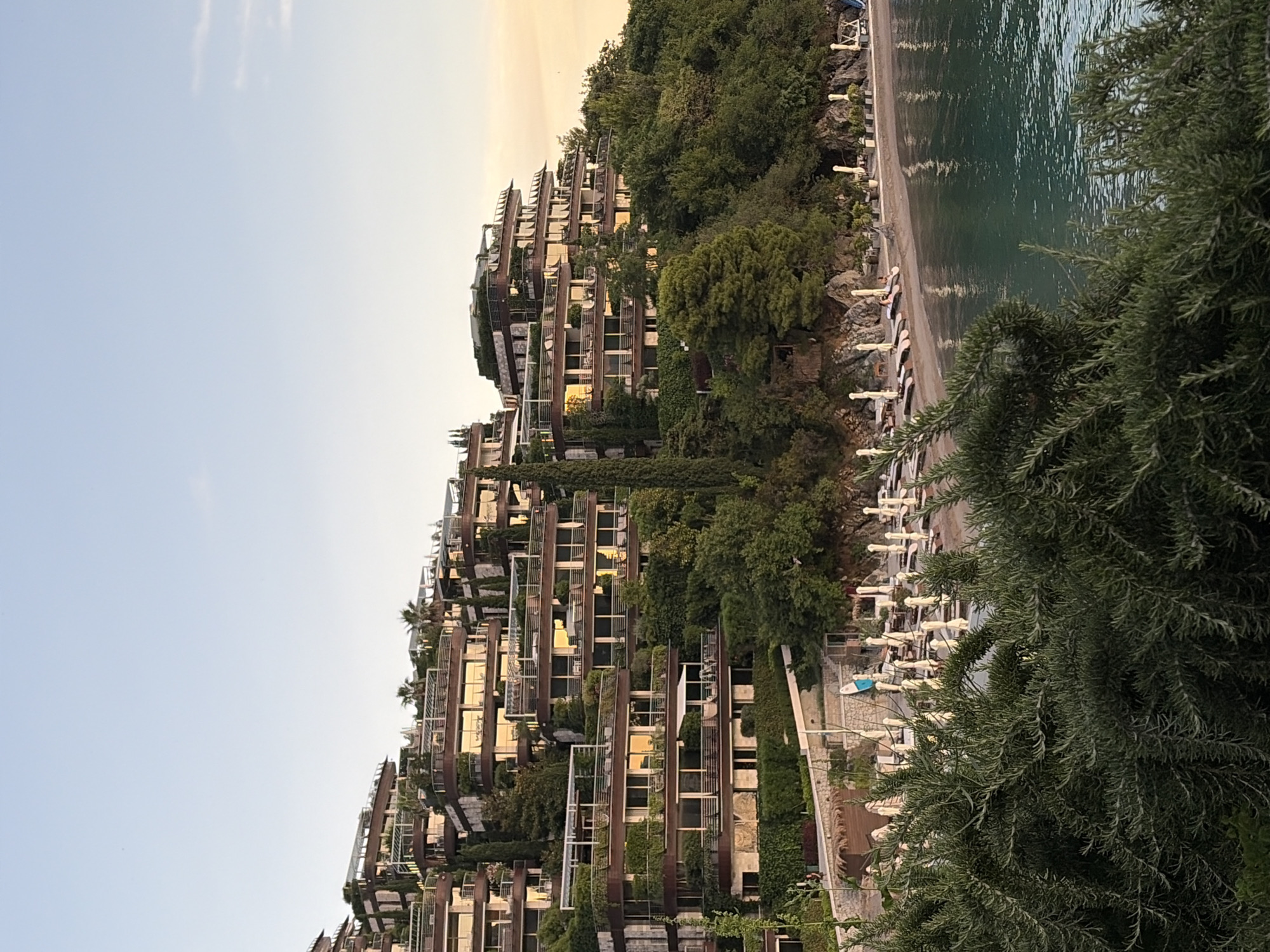 The image shows a hillside resort complex on the coast of Budva, Montenegro. The resort features several multi-story buildings with tiered architecture, extensively covered in greenery and featuring balconies and large windows. Below the resort, a beach area is visible with rows of lounge chairs and umbrellas. The sea is calm and has a turquoise hue. The sky is clear with a hint of sunset glow in the upper right corner. In the foreground, lush green foliage obscures a portion of the view. A few people are discernible on the resort's terraces and near the beach. The overall scene conveys a sense of luxury and a tropical coastal vacation setting.