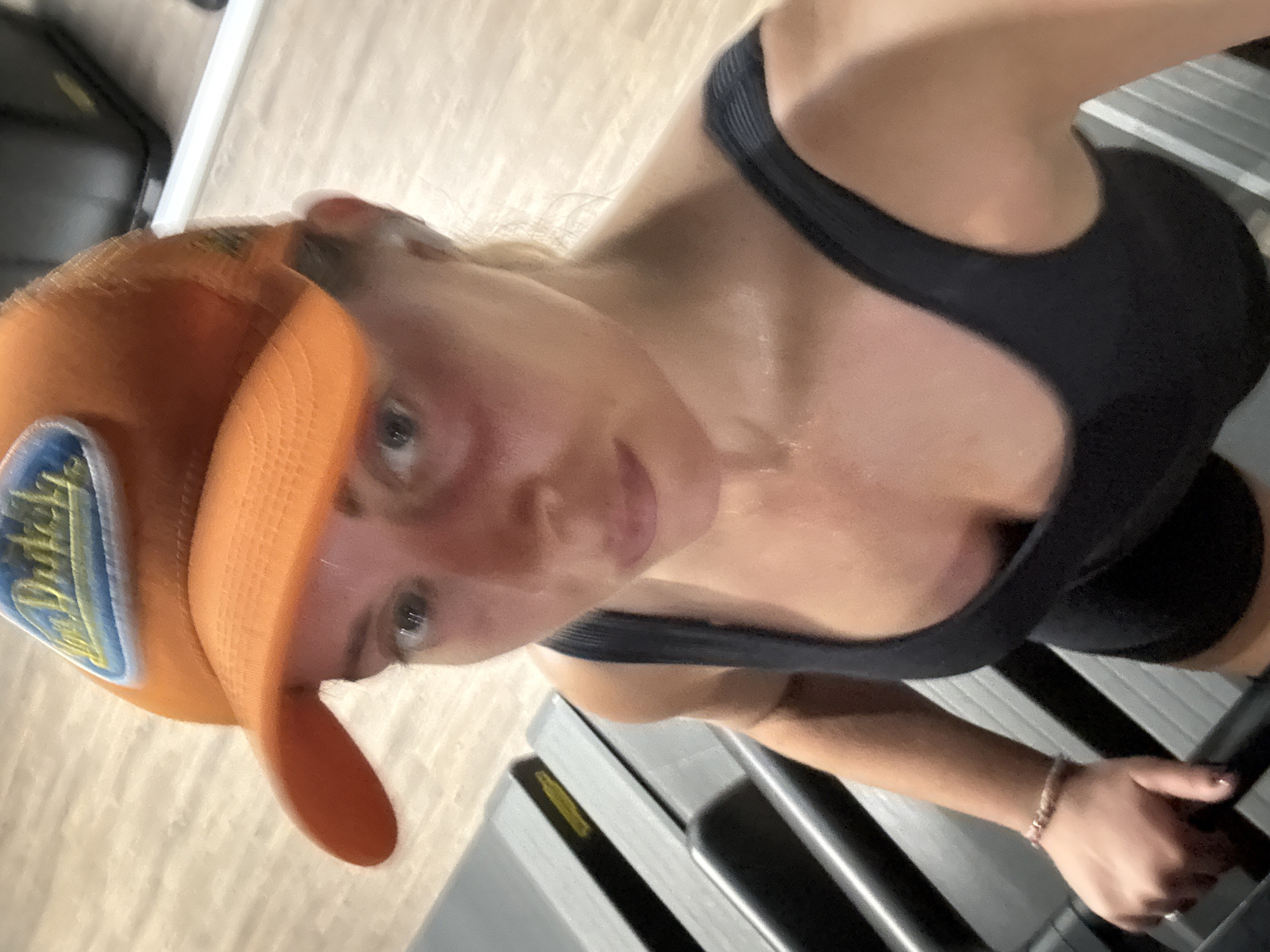 A fair-skinned woman wearing an orange baseball cap and a black sports bra is positioned on a treadmill. The treadmill is gray with black accents, and the woman's right hand is gripping the handle. The background consists of a neutral-toned floor. The woman appears to be perspiring. The image does not contain information about the location being in the city of Paris, France.