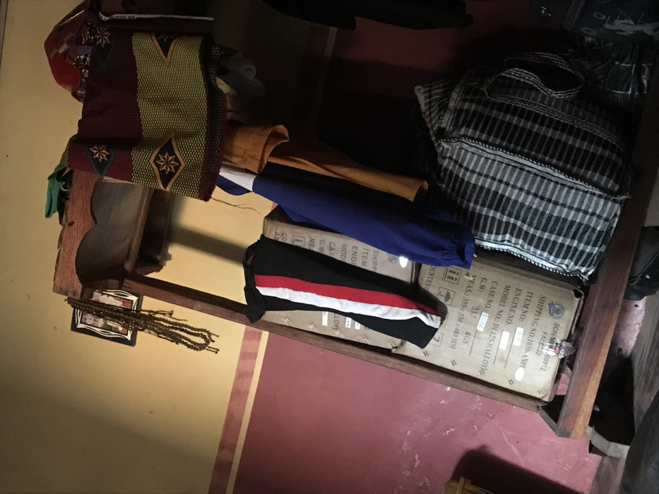 A cluttered wooden shelf with various items rests against a wall with a red and yellow color scheme. A woven, patterned vest, blue and yellow clothing, and a black and white striped item are stacked on top. The shelf also holds a picture frame with beads and sits on a box labeled "SON/MP 00912 FACE332" and other shipping details, including item and engine numbers. The box dimensions are listed as 355x330x480 mm.
