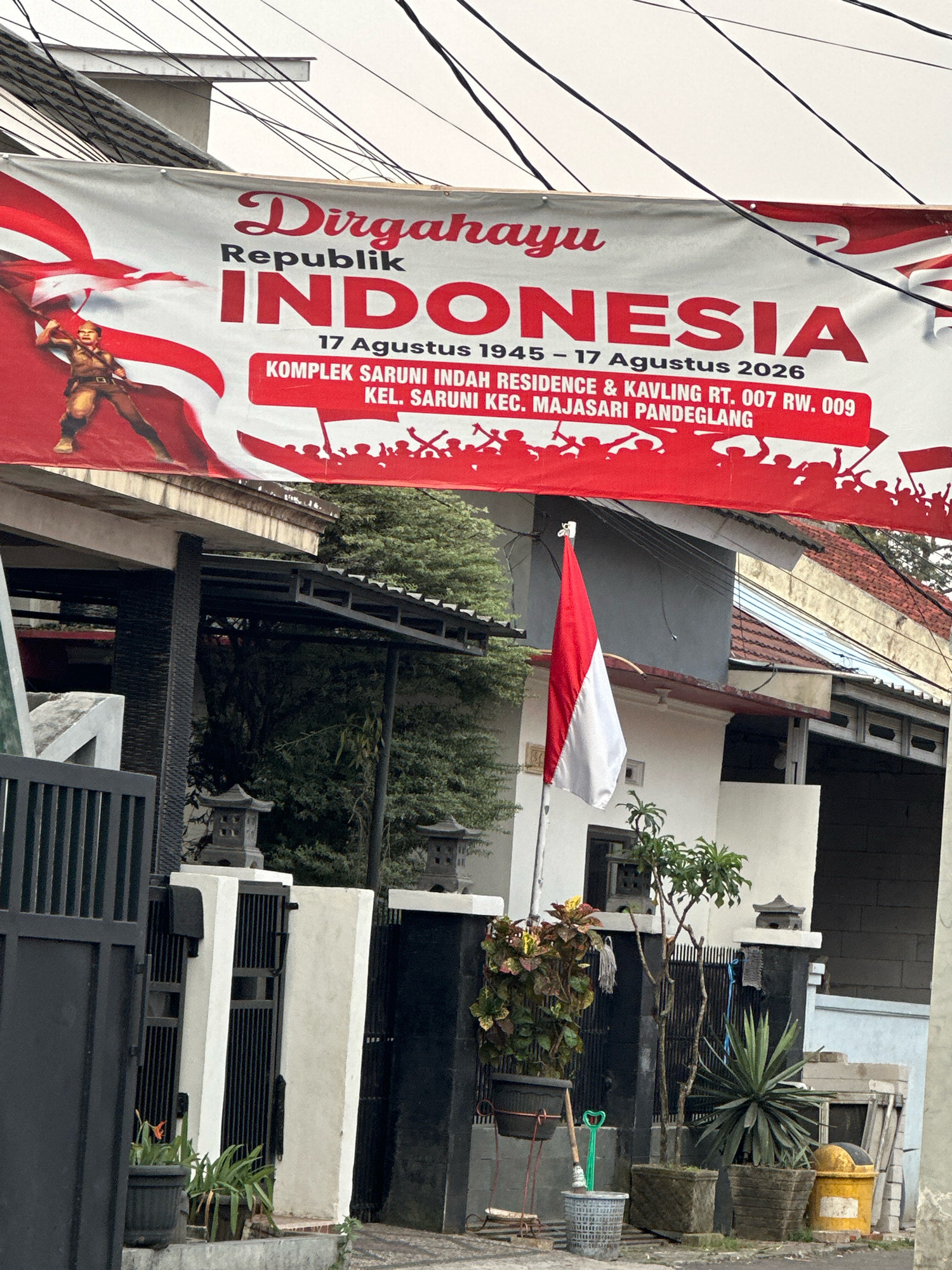 A red and white banner celebrating Indonesia's independence is displayed above a street in Saruni, Indonesia. The banner features text that reads "Dirgahayu Republik INDONESIA" and "17 Augustus 1945 - 17 Agustus 2026". Below this, it lists the location as "KOMPLEK SARUNI INDAH RESIDENCE & KAVLING RT. 007 RW. 009 KEL. SARUNI KEC. MAJASARI PANDEGLANG". The banner also depicts a stylized soldier raising a flag and a crowd of silhouettes. A small Indonesian flag is flying on a pole in front of a building. The surrounding environment includes houses, fences, trees, and electrical wires. The street appears to be paved and there are several potted plants visible.