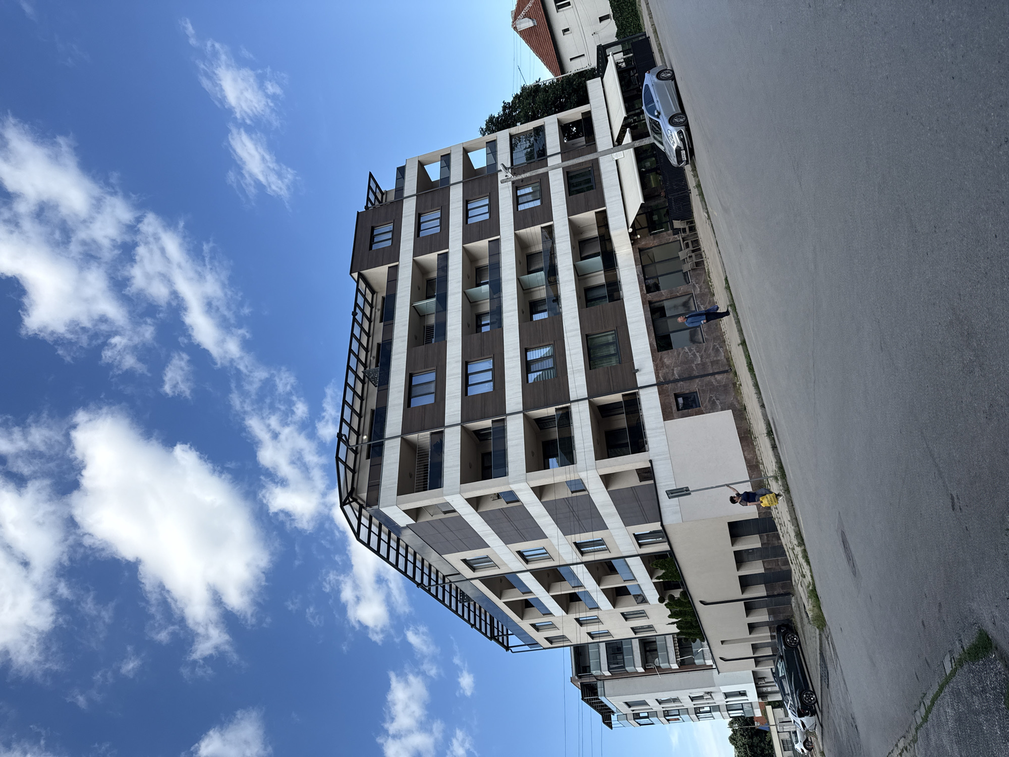 The image depicts a modern apartment building with a dark brown and light beige facade, featuring multiple balconies with glass railings. The building has a flat roof with an intricate metal structure. In front of the building, there is a street with a silver sedan parked. A person in a blue shirt and dark pants is standing on the sidewalk near the building entrance. The sky above is blue with scattered white clouds. To the right of the apartment building, a smaller, traditional-style house with a red-tiled roof is visible. The image appears to be taken in an urban setting in Vratsa, Bulgaria.