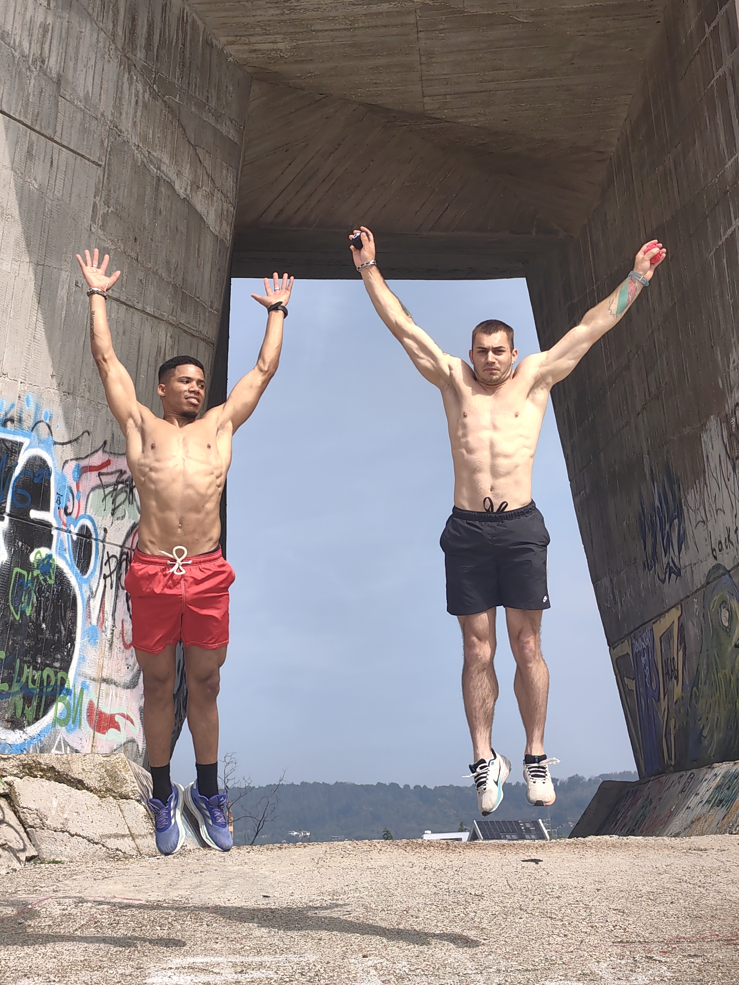 Two adult males are depicted mid-jump under a large concrete overpass or bridge structure, characteristic of the area near the Asparuhov Bridge in Varna, Bulgaria. Both subjects are shirtless, revealing athletic builds, and have their arms raised above their heads.
The male on the left, of apparent mixed ethnicity, wears red athletic shorts with a white drawstring and blue and purple athletic shoes over black socks. He has bracelets on both wrists and displays an upward gaze with a slight smile.
The male on the right, appearing Caucasian, wears black athletic shorts with a white Nike logo and light-colored athletic shoes. He exhibits a prominent green and blue tattoo on his right forearm, bracelets on both wrists, and holds a small red object in his raised right hand. His expression is neutral as he looks directly at the camera.
The imposing concrete structures framing the subjects are heavily weathered, displaying cracks and uneven textures, and are extensively covered with vibrant, multi-colored graffiti. The ground beneath the subjects is a light-colored, coarse concrete or paved surface with small aggregate. The background consists of a clear blue sky above distant hills or land formations. The scene is illuminated by bright daylight, casting distinct shadows beneath the jumping individuals.