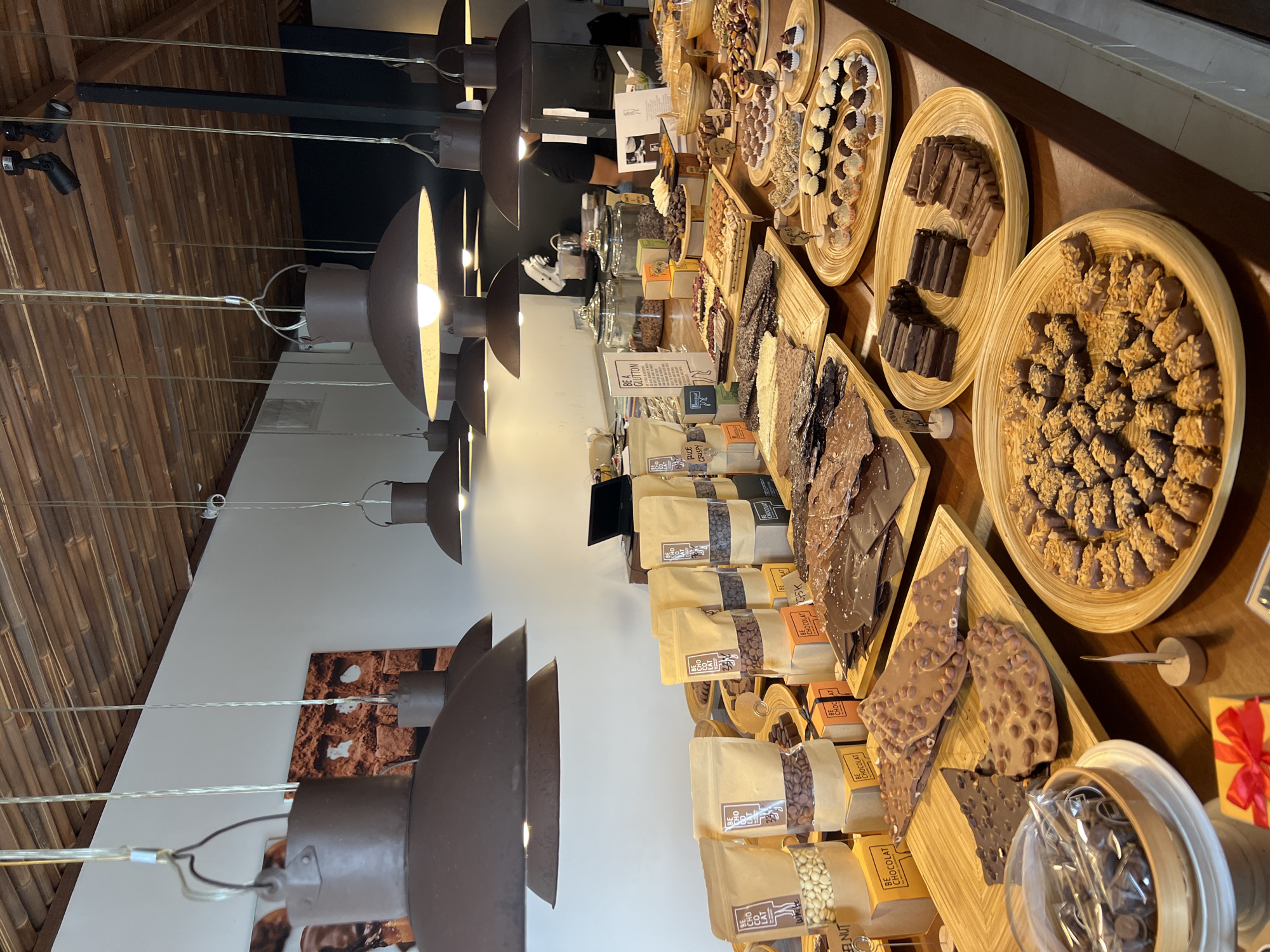 This image shows a retail display of "BE CHOCOLAT" products. The location appears to be an indoor shop, given the presence of lighting fixtures, a ceiling, and wall decorations.
The main subject is a long wooden counter with various chocolate products arranged for sale. The products include bags of chocolate, chocolate bars, chocolate bark, and small chocolate candies. The products appear to be packaged and presented in a way that suggests a gourmet or artisanal quality.
The environment is well-lit, with a combination of natural and artificial lighting. The store has modern, minimalist decor, with white walls and a light-colored wooden ceiling. A rack with overhead lamps gives light, and the wall on the left has a brown rectangular piece with patterns.
The image does not contain people.
