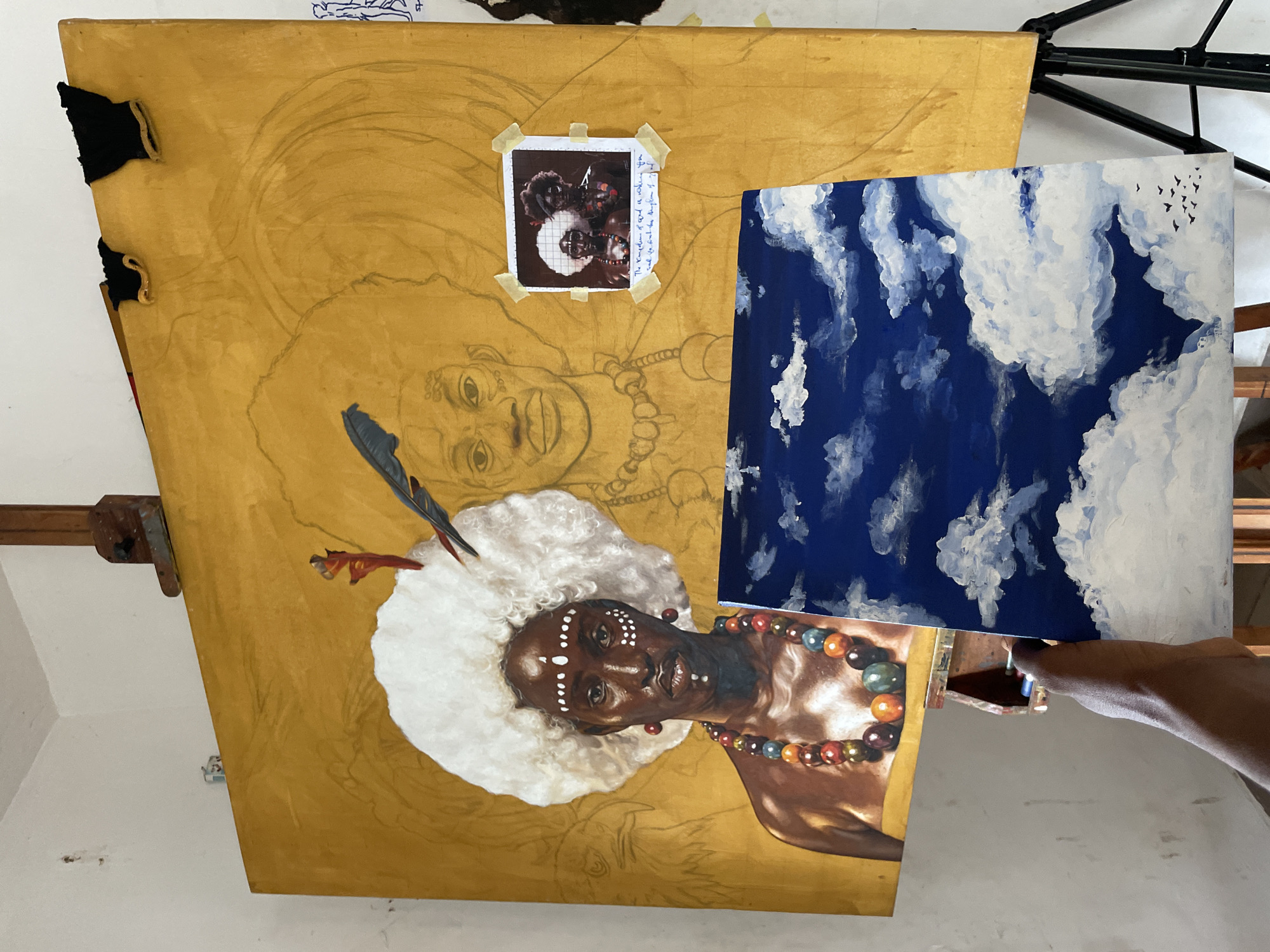 An artist's studio in Ado, Nigeria, is depicted with two paintings in progress and a reference image. The central focus is a large, vertically oriented painting on an easel, featuring a partially completed portrait of an African woman on a golden-yellow background. The completed section shows the woman's head and upper torso, adorned with white face paint dots, a fluffy white headpiece with a feather, and a vibrant, multi-colored beaded necklace. Her skin is rendered in rich, warm tones. Below this, a pencil sketch of another woman's face and upper body, with an afro-like hairstyle and a beaded necklace, is visible, indicating an underpainting or a second figure in the composition.
Taped to the upper part of the large painting is a small, polaroid-style reference photograph showing a similar subject, possibly the same woman, overlaid with a grid pattern. Beside this photo, handwritten text reads: "The Kingdom of God is within you. Seek ye first the Kingdom of God".
To the right, a hand holds a smaller, separate painting depicting a deep blue sky filled with fluffy white clouds and a scattering of small, dark bird silhouettes in the upper right. This smaller canvas appears to be a study or a component for a larger work.
The large painting is secured to an unseen support by black clips on its top-left edge. A black metal easel is partially visible behind the canvases, suggesting an active workspace. The overall scene captures a moment of artistic creation, blending portraiture with celestial elements and personal reflection.