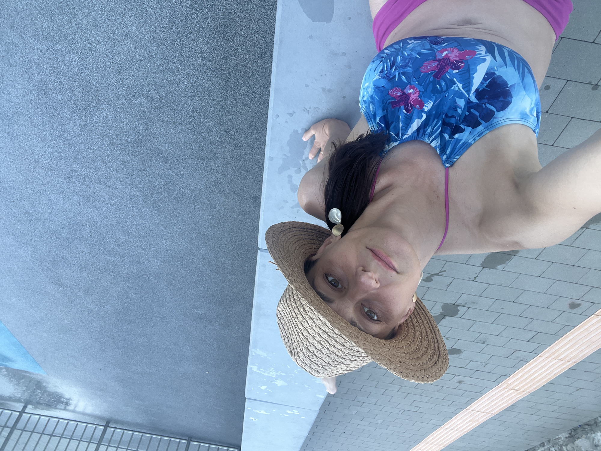 A woman wearing a straw hat and a floral bikini poses for a selfie by the edge of a pool. She is sitting on a concrete ledge, with her left arm resting on the ledge. The background shows a paved area with yellow tactile paving and the pool surface, which is wet and reflects the sky. The woman has brown hair and is looking at the camera with a slight smile. She is wearing large, round, gold earrings. The image is taken from a high angle.