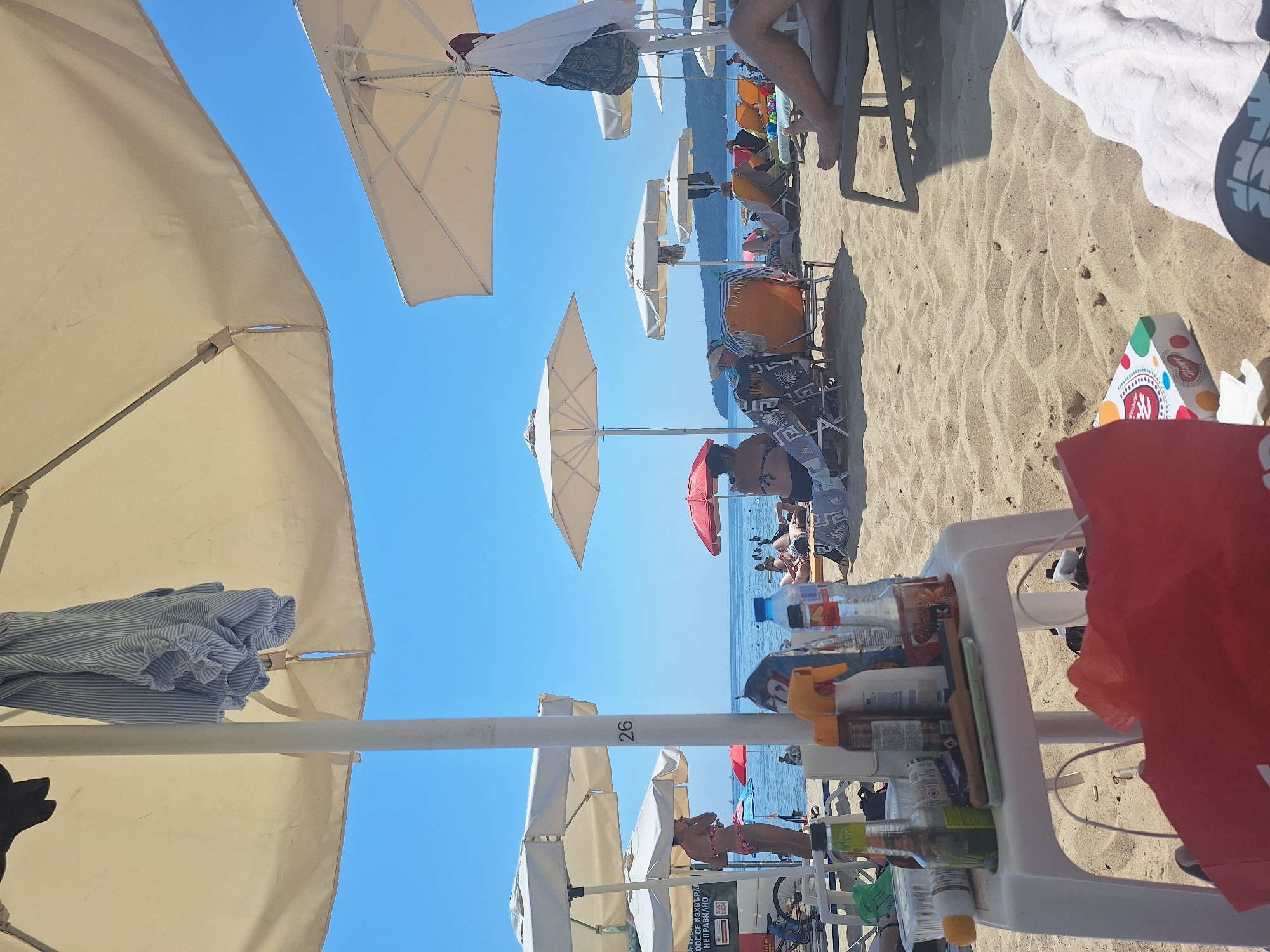 The image depicts a crowded beach on a sunny day, likely in Varna, Bulgaria. Numerous beach umbrellas are set up, providing shade for sunbathers lounging on chairs and towels. The sand is a prominent feature, with ripples and footprints visible. In the foreground, a white plastic table holds several items, including bottles of drinks, a spray bottle, and what appears to be snacks in a box. Beyond the immediate foreground, people are scattered across the beach and in the shallow water. The sea is calm and blue, meeting the horizon under a clear sky. In the background, a distant coastline with trees can be seen. A white and blue striped shirt is hanging on a pole, presumably a beach umbrella pole, in the upper left of the image. The number "26" is visible on a white pole in the middle ground.