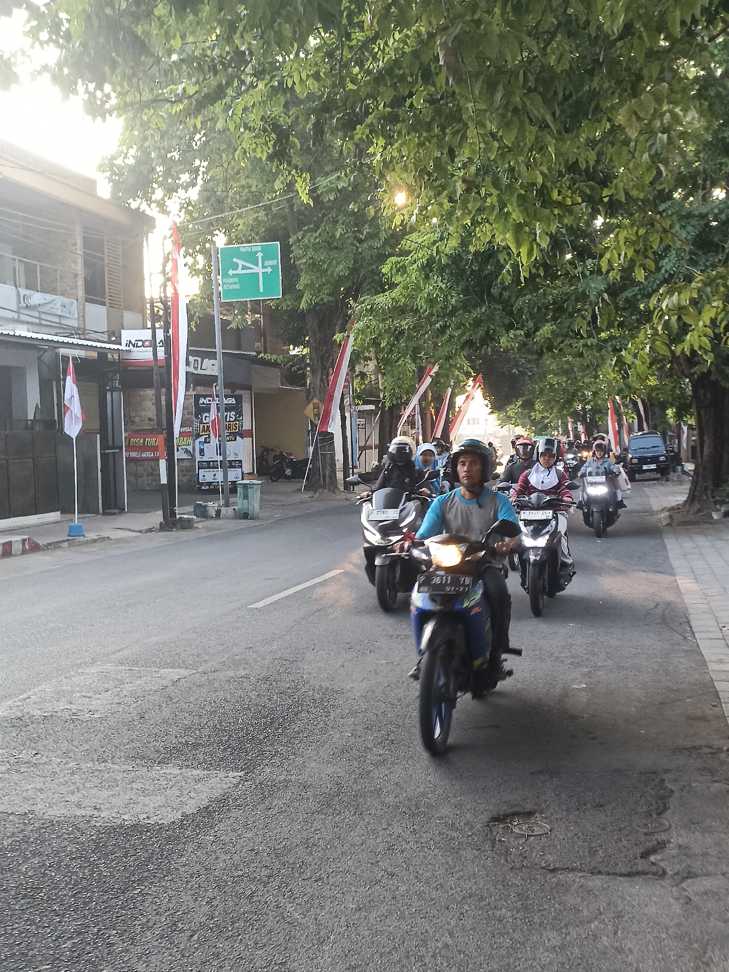 The image captures a street scene in Banyuwangi, Indonesia, likely during daylight hours given the bright sunlit foliage and the shadows cast. A procession of motorcycles, ridden by individuals wearing helmets, travels down the asphalt road. The motorcycles appear to be a mix of automatic and manual transmission models. Many of the riders are wearing casual clothing, and some passengers are visible on the back of some motorcycles. The road itself shows some signs of wear, with patches and a manhole cover visible.
Flanking the road are tall, leafy trees that create a canopy over parts of the scene. On the left side of the road, there are buildings with storefronts. One prominent sign reads "INDO G". Another sign appears to advertise "GRATIS AKSESORIES". Indonesian flags, red and white, are displayed vertically along the street, suggesting a national holiday or celebration. A green directional signpost is visible, indicating routes to "PANTAI" (beach) and "JEMBER". On the right side of the road, a paved sidewalk runs alongside the street, with a parked dark-colored vehicle further down the road. The overall atmosphere suggests a typical urban street in Indonesia, possibly on a route leading to a popular destination like a beach, given the signage.