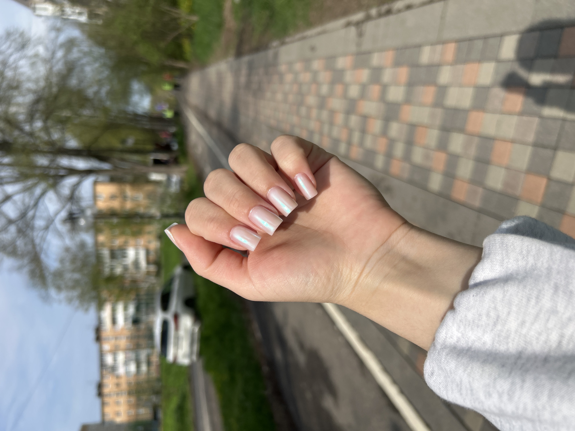 A vertical outdoor shot depicts a hand with iridescent square-shaped nails. The hand is positioned in the foreground, slightly angled towards the right. The background shows a paved pathway with a shadow figure, likely the photographer, on the right. To the left, there are trees, a white car, and a multi-story building. This location is in Klin, Russia.