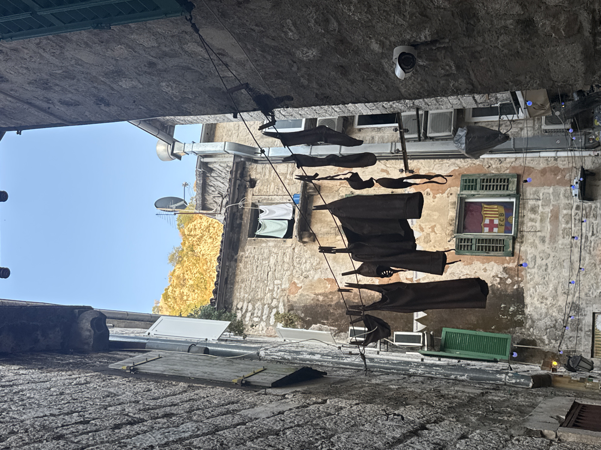 The image depicts a narrow street in Kotor, Montenegro, characterized by ancient stone buildings and laundry hanging on a clothesline. The perspective is from ground level, looking upwards into the sky, which is clear and blue. On the left, a rough-hewn stone wall rises steeply, partially obscured by a vertical white panel and a section of grey metallic siding. A green-shuttered window is visible further up the wall.
Dominating the center of the frame is a clothesline strung across the alleyway, laden with various articles of dark-colored clothing, including what appear to be dresses or tunics, and possibly underwear. The clothing is secured by clothespins.
On the right side of the alley, another stone building stands, this one with a rougher texture. A white security camera is mounted on this wall, facing downwards. Further up this building, a window is visible, fitted with green shutters, and behind the glass, a flag with a red and yellow crest, possibly that of FC Barcelona, is displayed.
In the background, more stone buildings are visible, some with exposed brickwork and weathered plaster. A hillside covered in yellow foliage can be seen at the very top of the frame. The overall scene suggests a residential area within the historic old town of Kotor, where everyday life unfolds amidst the backdrop of centuries-old architecture.