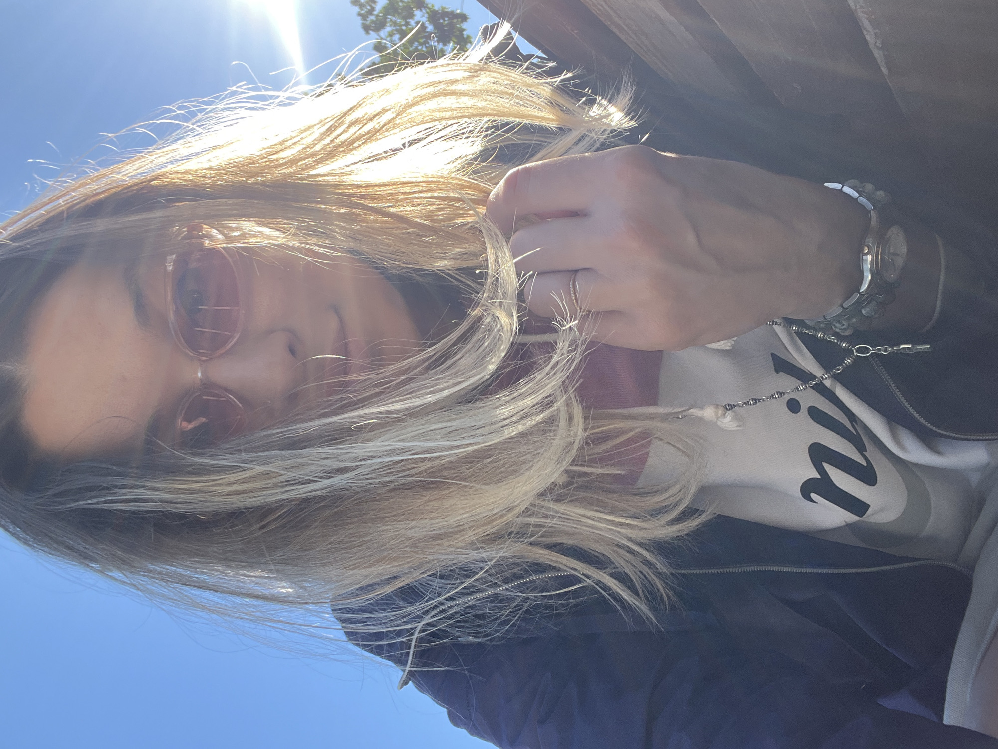A woman with blonde hair and pink sunglasses is photographed from a low angle, with the sky as the background. She is wearing a navy blue jacket over a white hoodie with a Nike logo. A silver necklace with a crucifix charm is visible, as well as a watch with a metallic band on her left wrist. Her hand is resting on her chest, and her hair is blowing in the wind. The scene appears to be outdoors, possibly in a park or an open area with some trees visible in the distance. The sun is shining brightly, creating lens flare in the image.