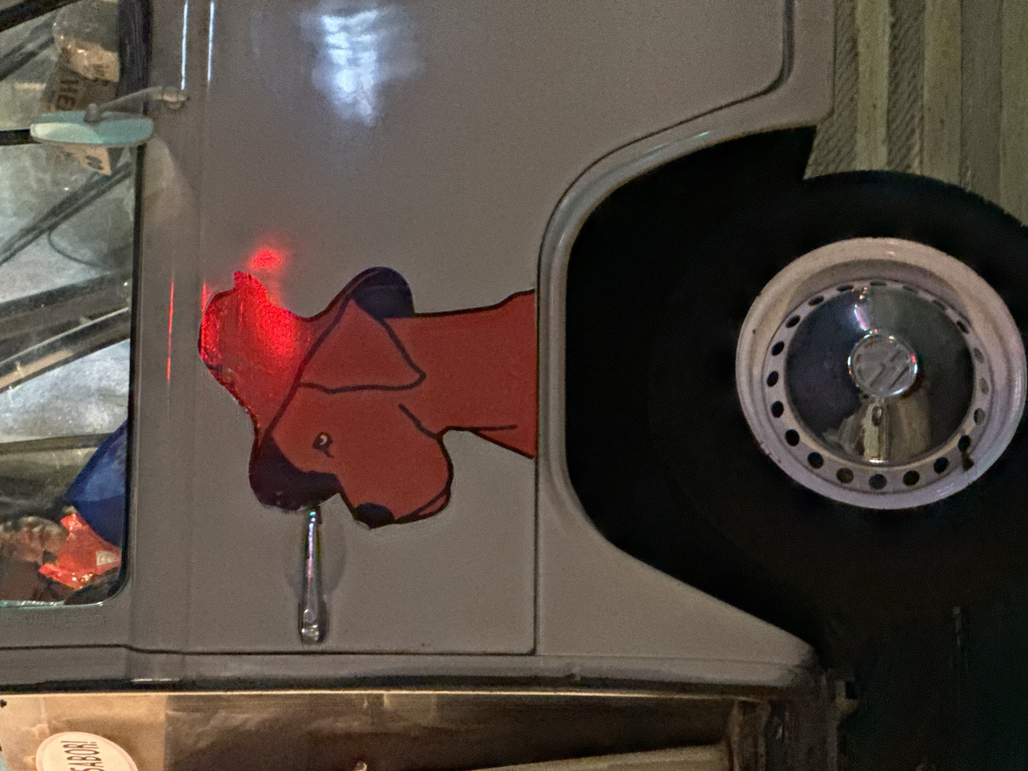 The image shows the side of a white Volkswagen van. On the van's panel, there is a cartoon-style drawing of a red dog wearing a hat. A partially visible car wheel, with a Volkswagen logo on the hubcap, is positioned beside the panel. The door handle is visible. There is a reflection in the window. At the bottom of the image, part of a sticker with the word "SABOR!" is visible. The setting appears to be Puebla, Mexico, due to the presence of Volkswagen, where its car assembly is located.