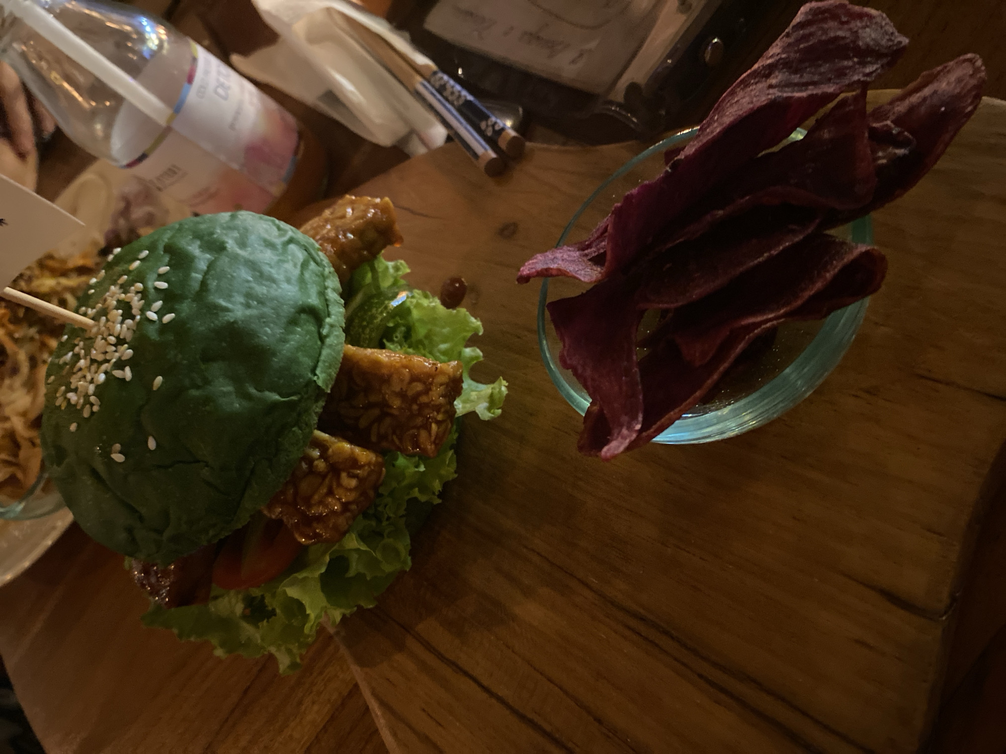 The image captures a culinary scene, possibly within a restaurant in Ubud District, Indonesia. A green-colored burger, adorned with sesame seeds, lettuce, tomato, and what appears to be tempeh is prominently displayed. Beside the burger, there is a glass bowl filled with dark purple chips, likely purple sweet potato chips. In the background, there is a water bottle and chopsticks. The entire scene is set on a wooden table. The lighting is subdued, suggesting a dim ambiance.