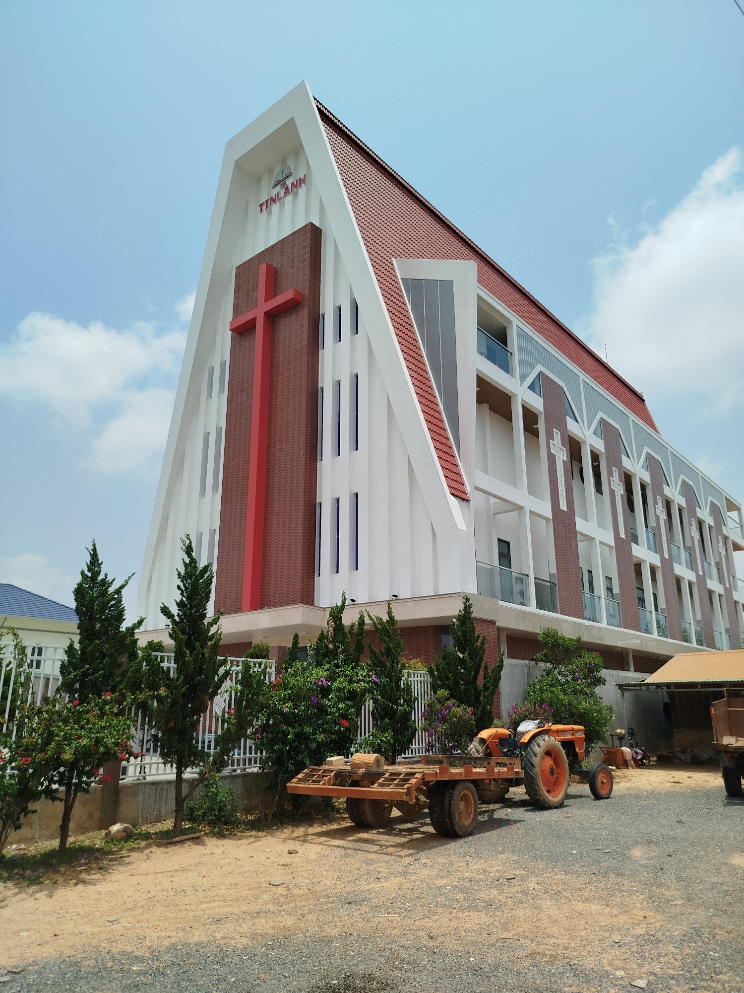 The image shows the Tin Lành Church in Đa Nghịt, Vietnam. The church is a multi-story building with a prominent A-frame facade, featuring a large red cross and the inscription "TINLANH" at the top. The building has a red tiled roof and white and brick-colored vertical sections. Adjacent to the church is a small residential building, and in the foreground, there is an orange tractor pulling a trailer loaded with logs. The surrounding area consists of gravel and dirt, and some greenery is visible near a white fence.