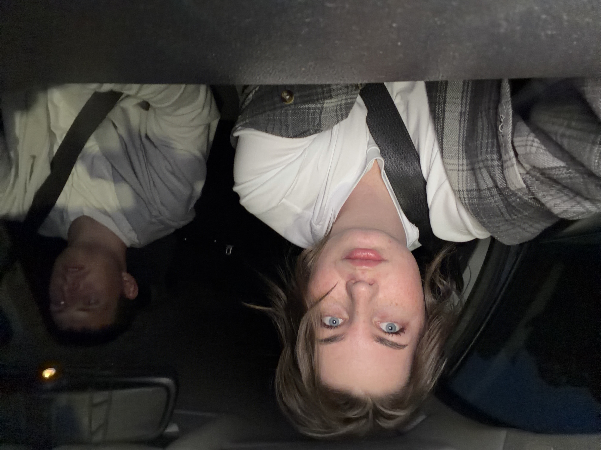 Two individuals are inside a car, both wearing seatbelts. The person on the right has blue eyes, light skin with freckles, and is wearing a white top with a gray plaid jacket. The person on the left is wearing a white shirt. The image appears to be taken from a low angle, looking up towards the individuals. The setting is inside a car at night. The location, Chaikovsky, Russia, cannot be confirmed from the given visual information.